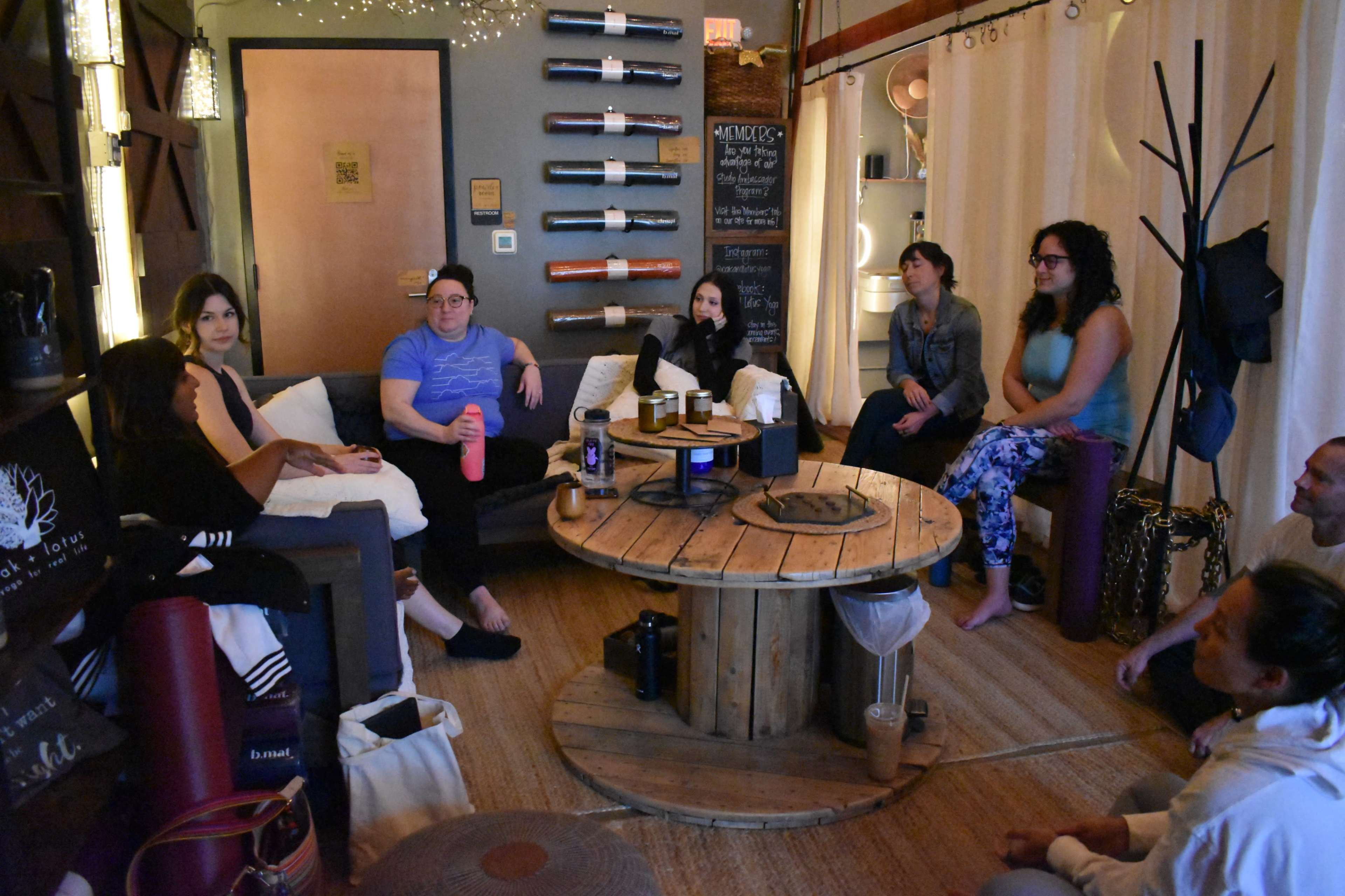 A group of individuals is seated in a cozy room, engaged in a discussion around a round wooden table, with yoga mats and various equipment visible in the background.