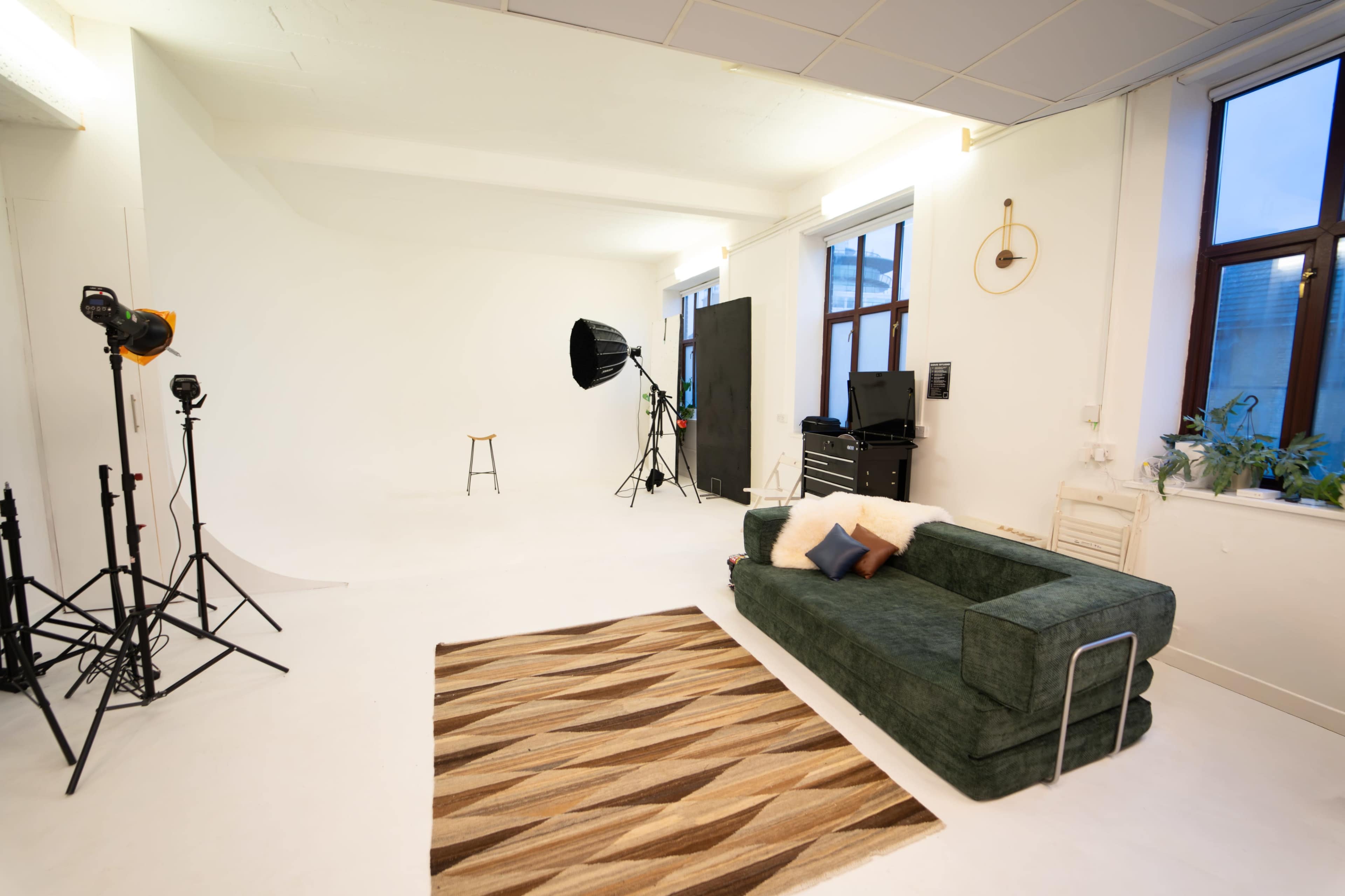 The image shows a photo studio with a green couch, a patterned rug, a stool, and various lighting equipment set up against a white backdrop.
