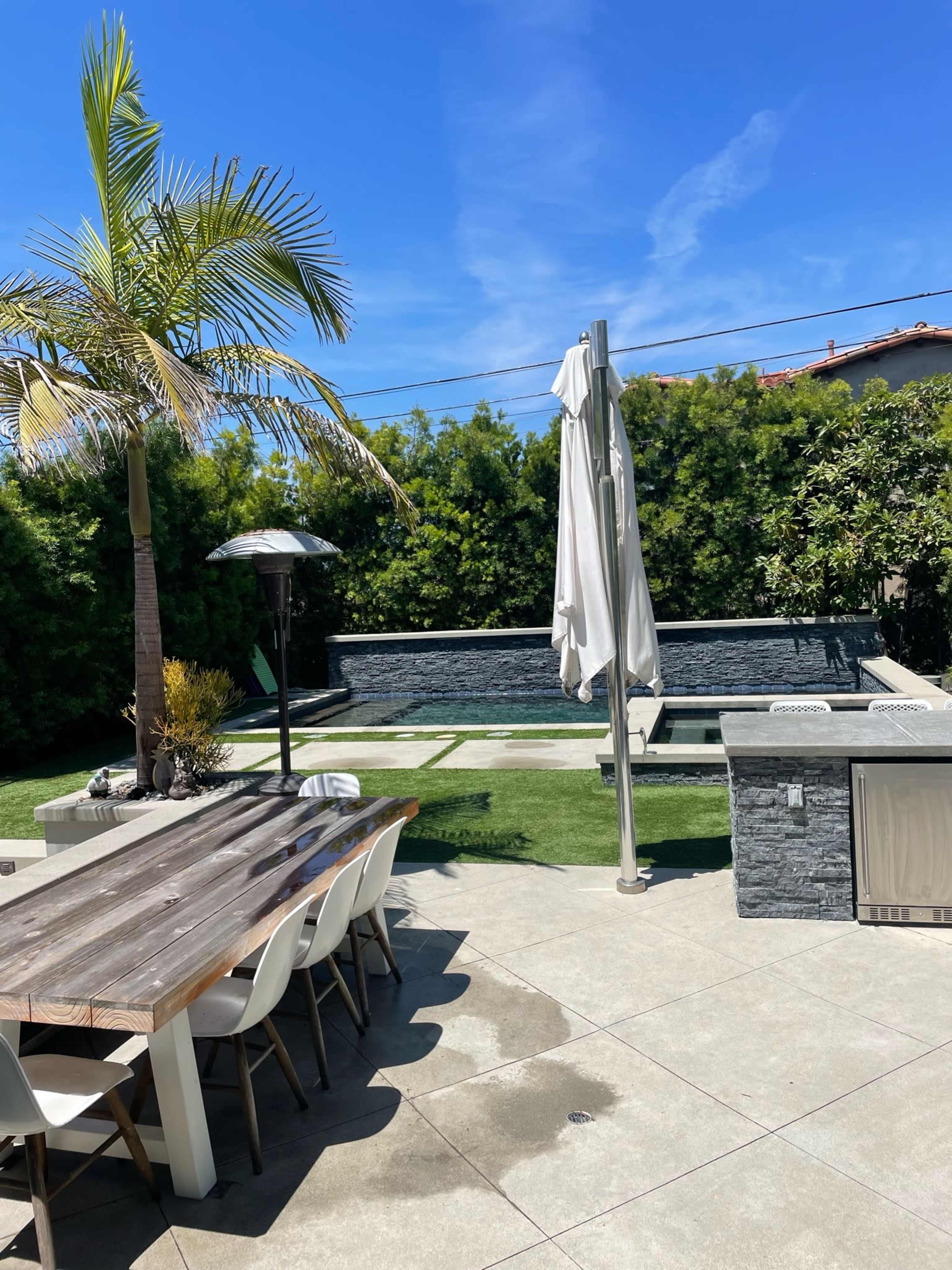 A backyard scene features a dining table and chairs, a pool surrounded by greenery, and a palm tree under a clear blue sky.