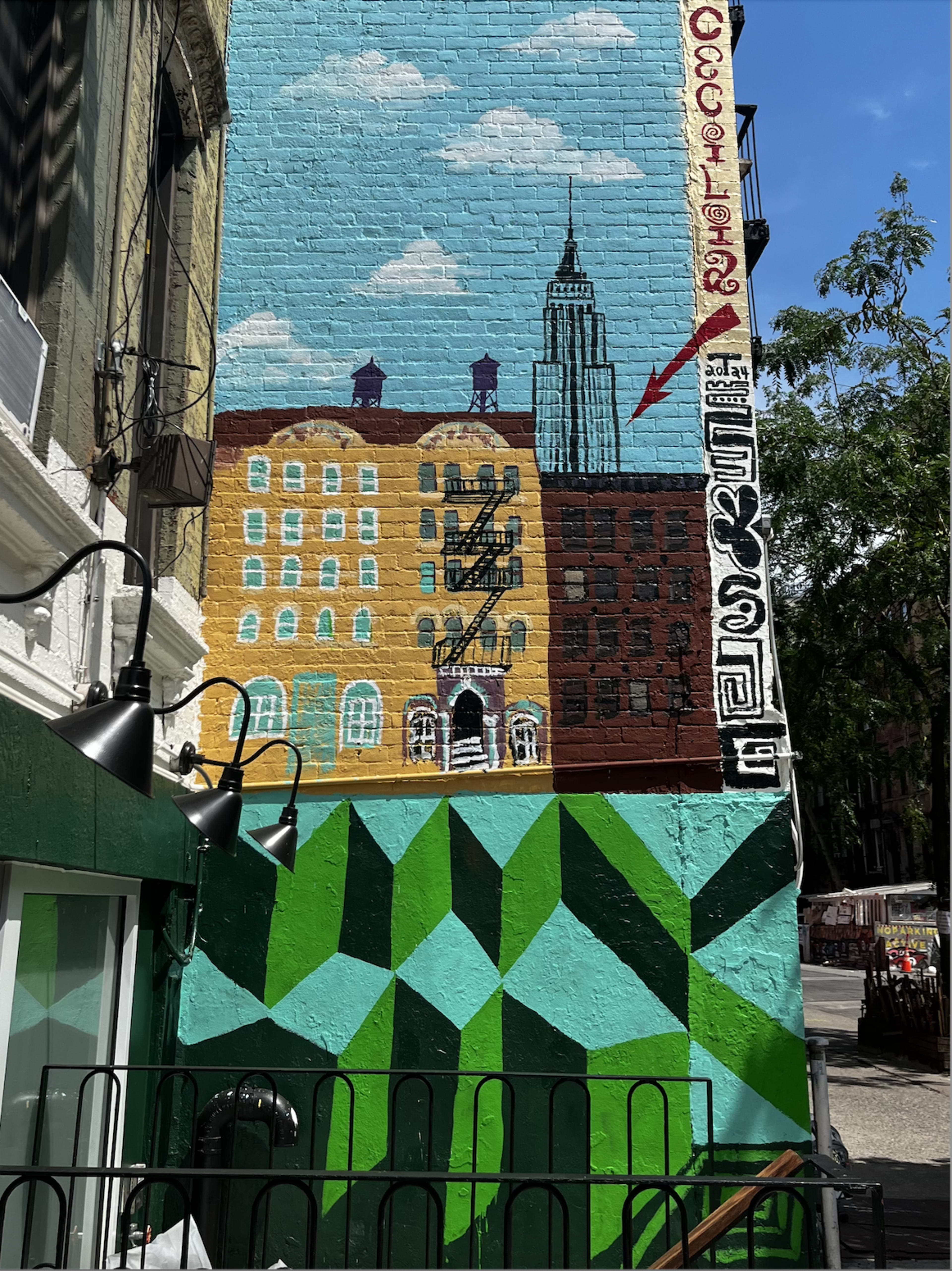 A colorful mural on a brick wall featuring buildings, an arrow pointing to the Empire State Building, and geometric green patterns below.