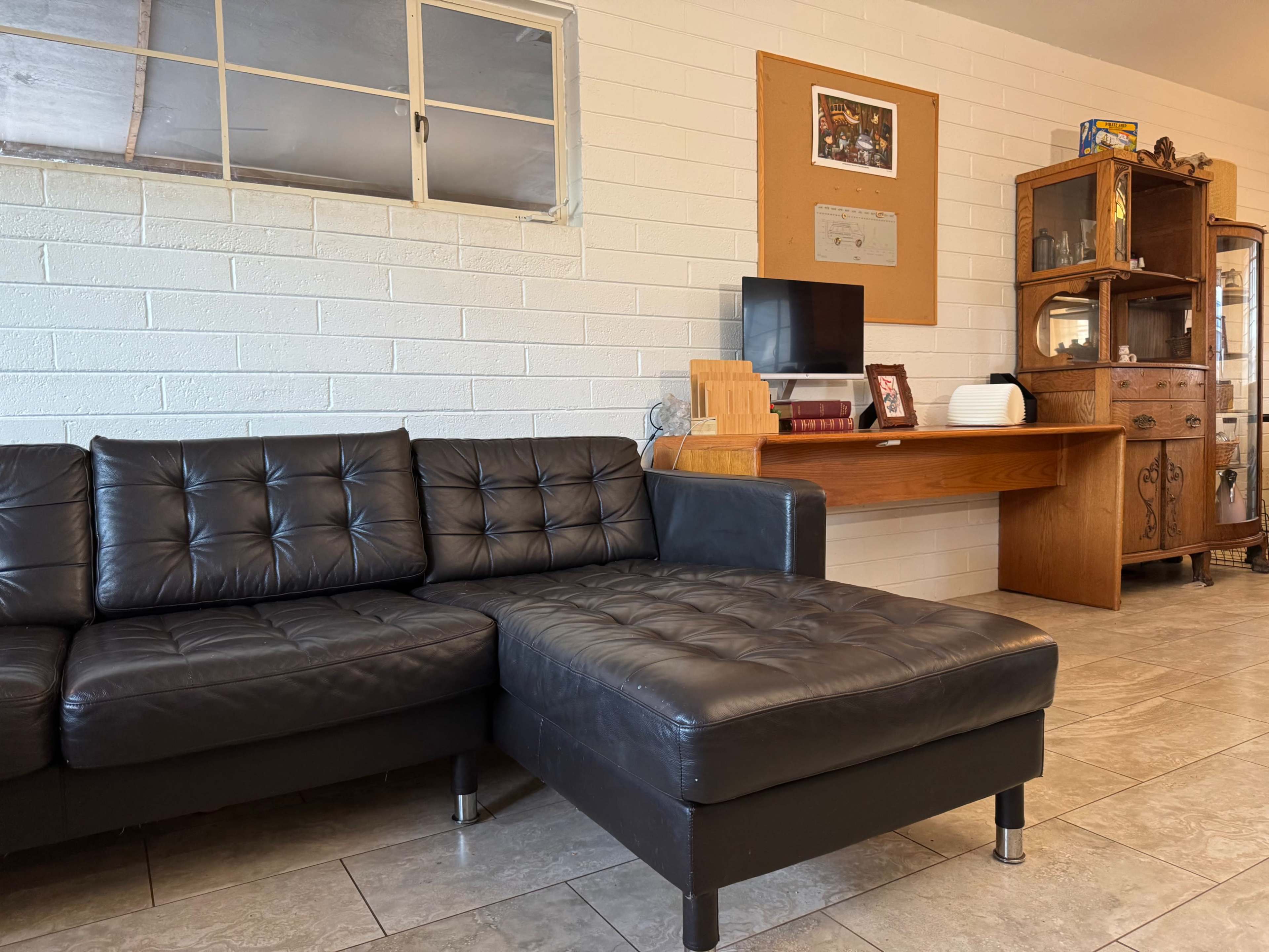 The image shows a black leather sectional couch in a room with a white brick wall, a wooden desk, and various wooden furniture pieces in the background.