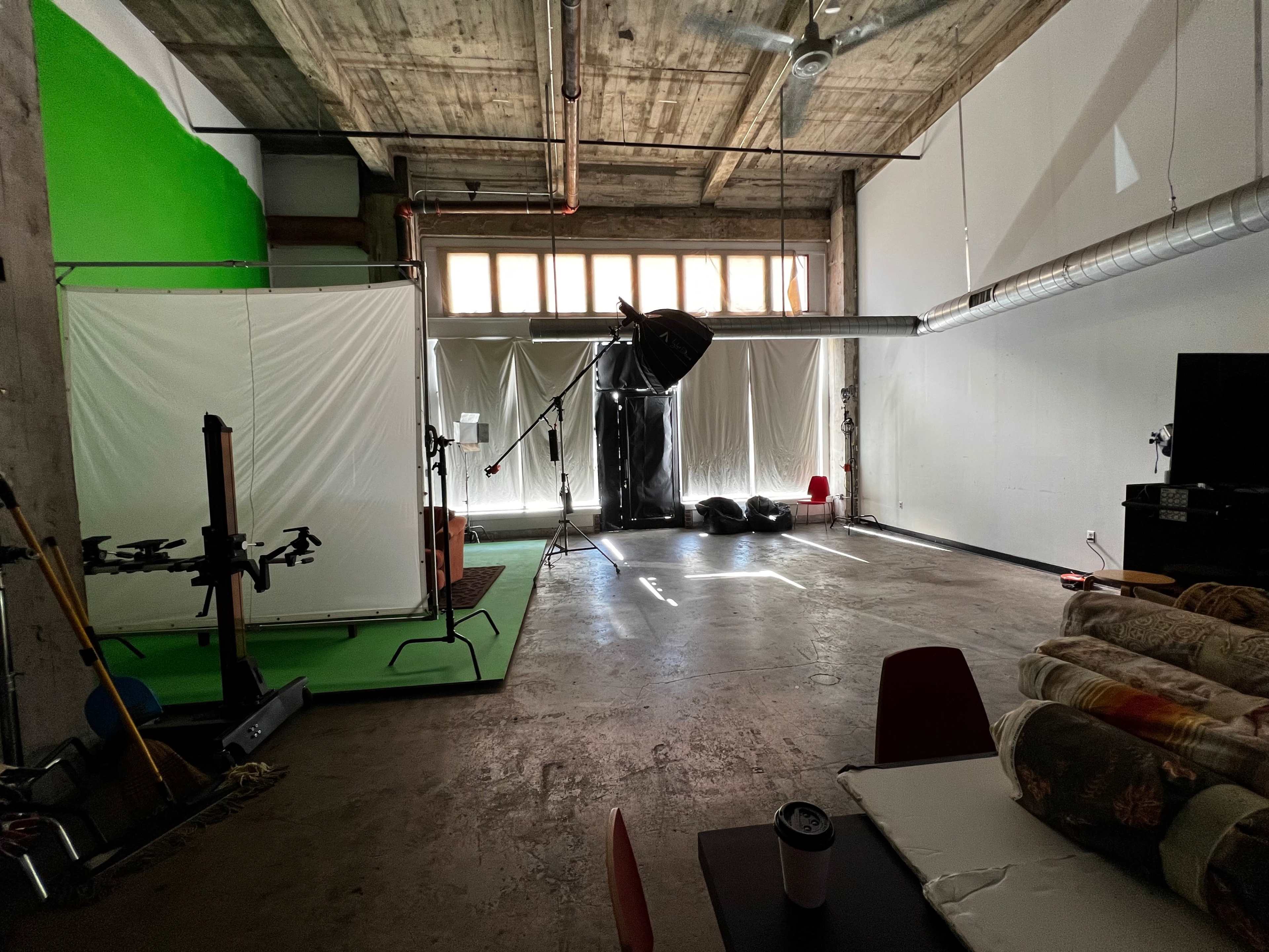 The image shows a studio space with green and white backdrops, various photography equipment, and natural light coming through large windows.