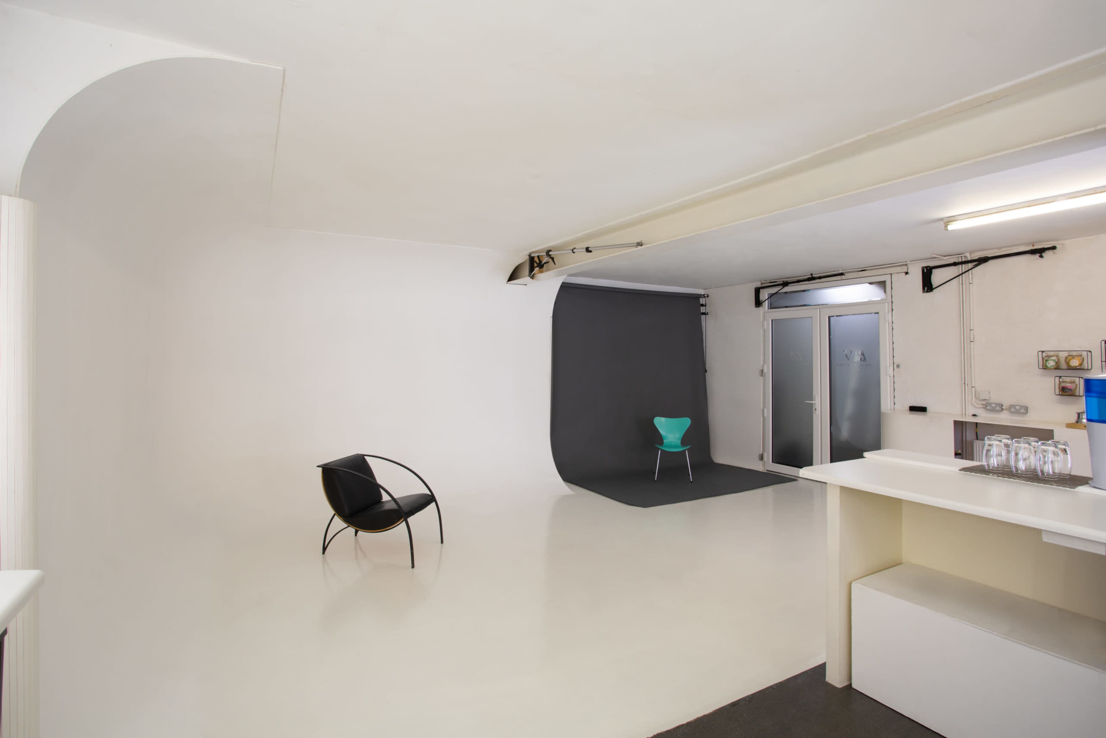 A spacious, minimalist studio with a white floor and walls, featuring a black chair in the foreground and a green chair on a gray backdrop in the background.