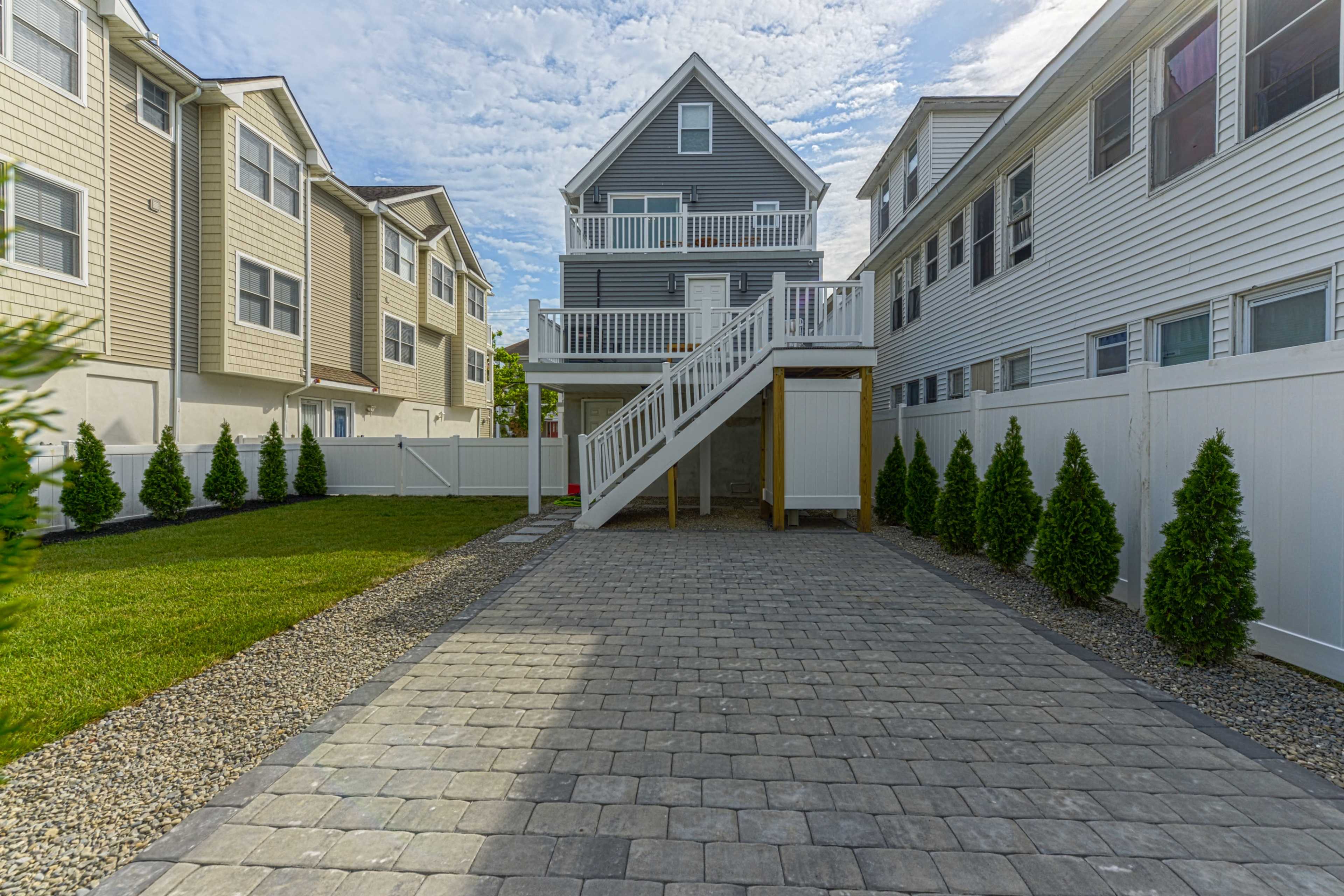 A two-story gray house with a front porch is situated in a paved backyard surrounded by decorative shrubs and adjacent homes.