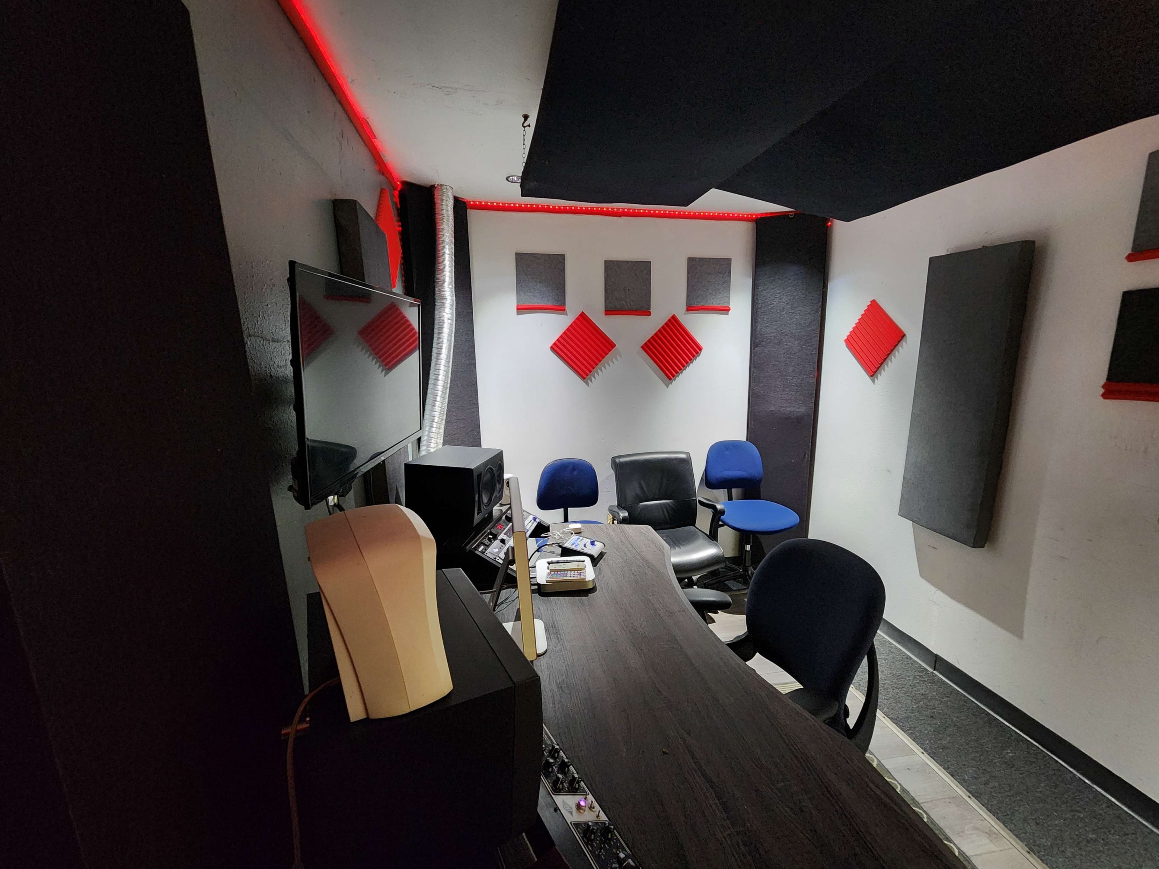 A modern recording studio with a large table, a television mounted on the wall, and soundproofing panels arranged in a geometric pattern.