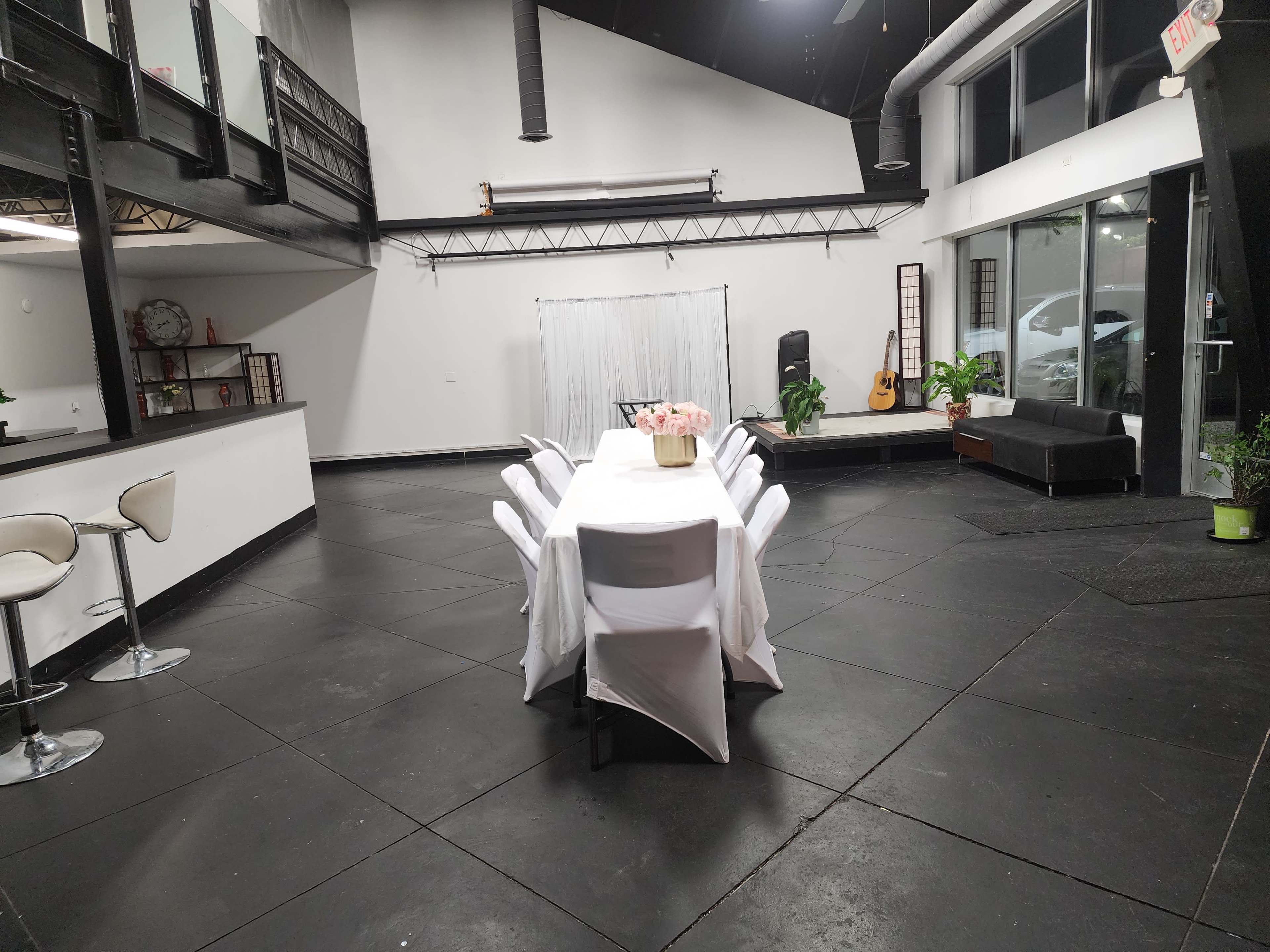 A spacious room with a long, white table set for dining, surrounded by chairs, and featuring a small stage or platform with a guitar in the corner.