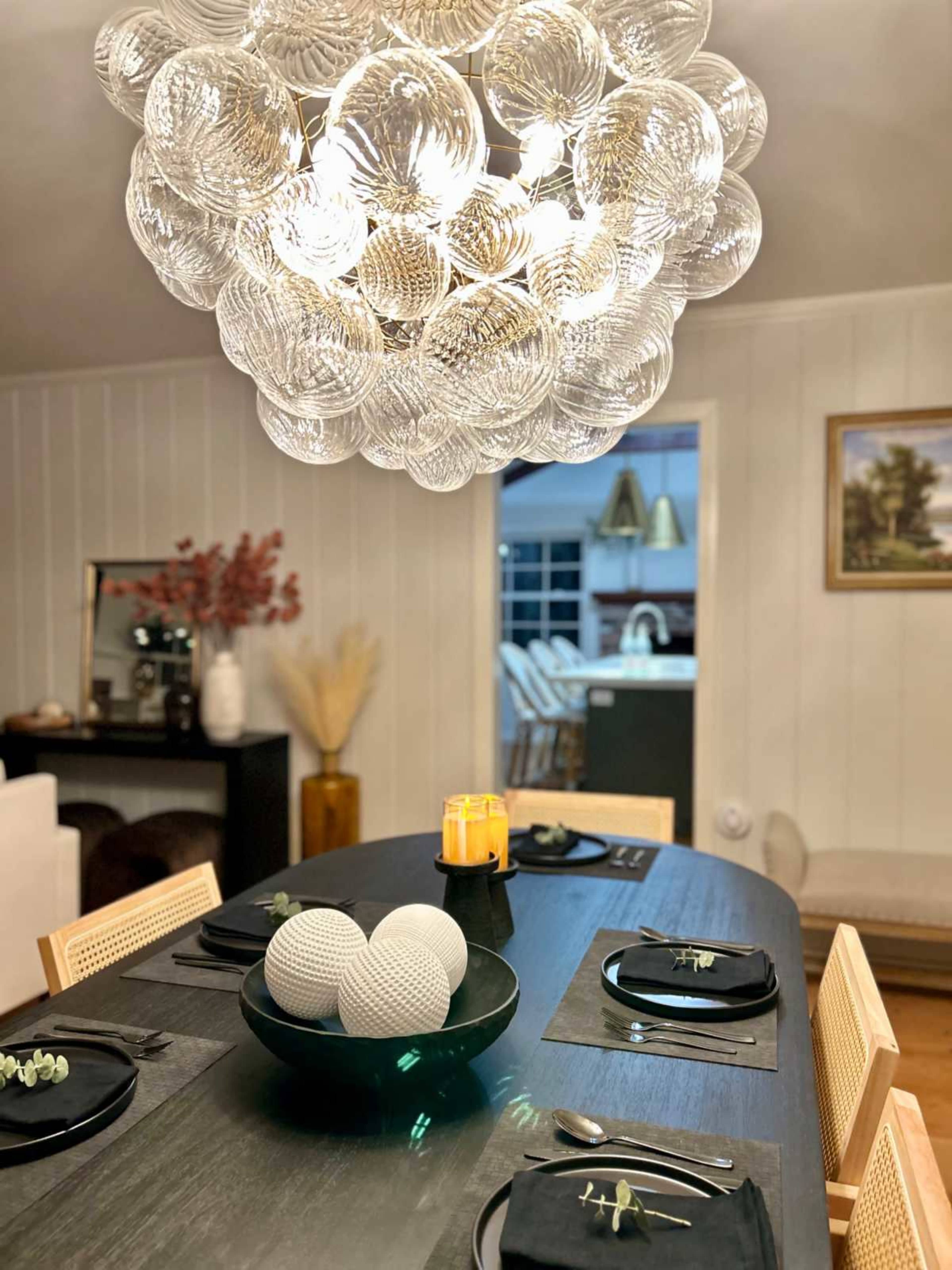 A modern dining room features a large circular light fixture above a dark wooden table set with black placemats, a bowl of decorative spheres, and a lit candle.