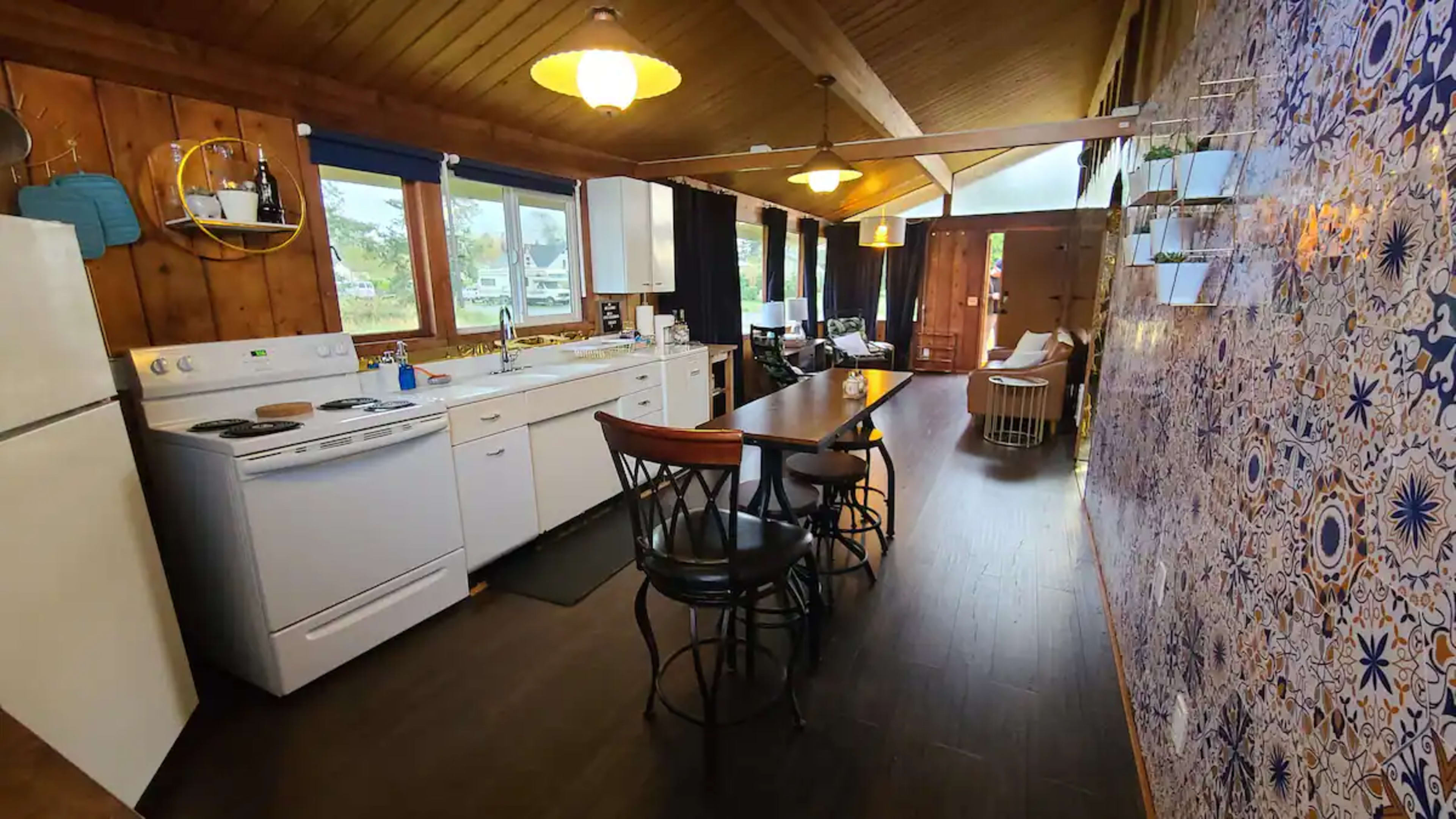 The image shows a modern kitchen with white appliances, a wooden dining table, and a decorative tiled wall, illuminated by pendant lights.
