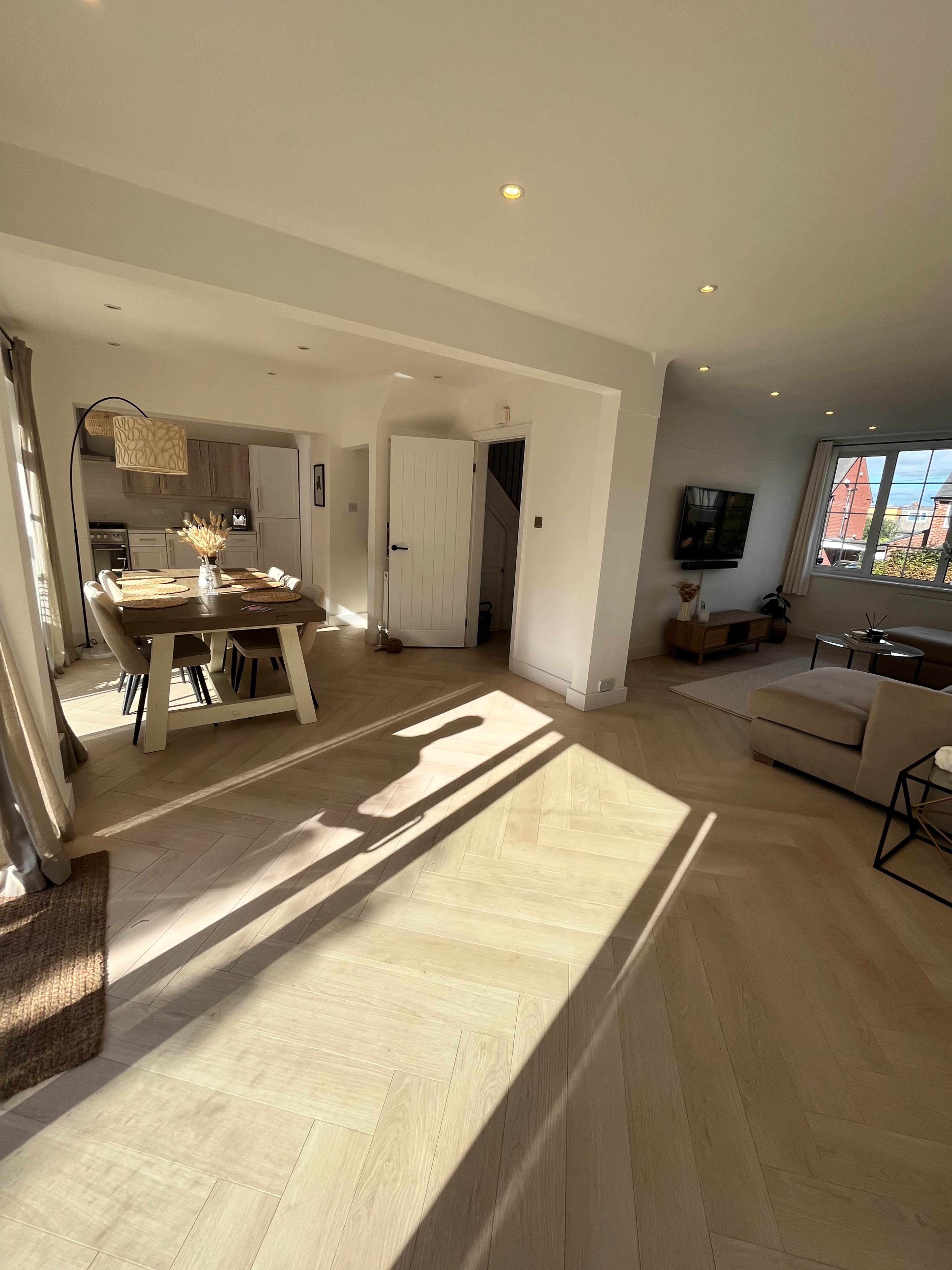 A bright, open living space with a dining area and a kitchen visible in the background, featuring wooden flooring and large windows.