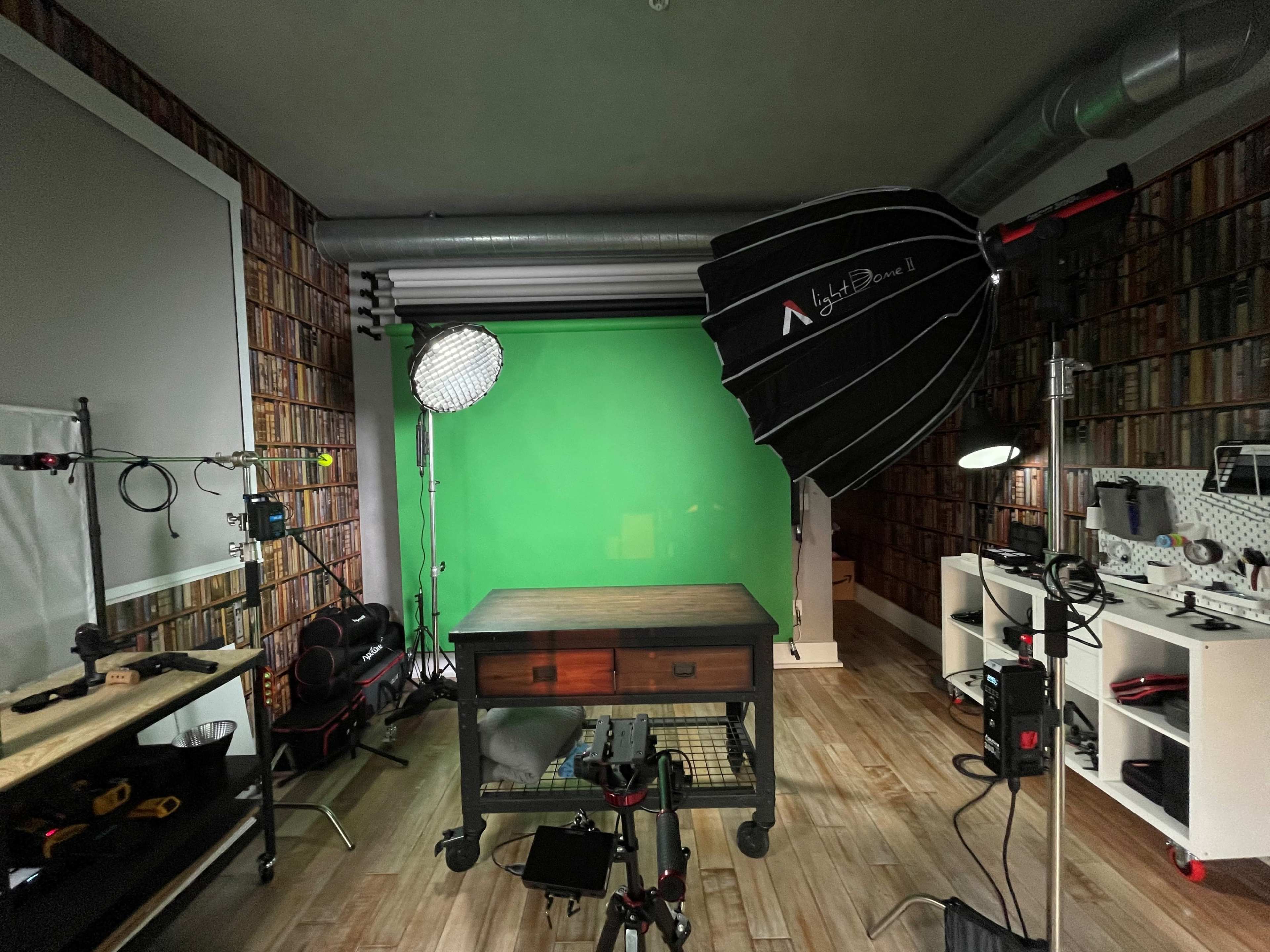 The image shows a well-equipped studio featuring a green screen backdrop, various lighting setups, and a central wooden table surrounded by photography and video equipment.