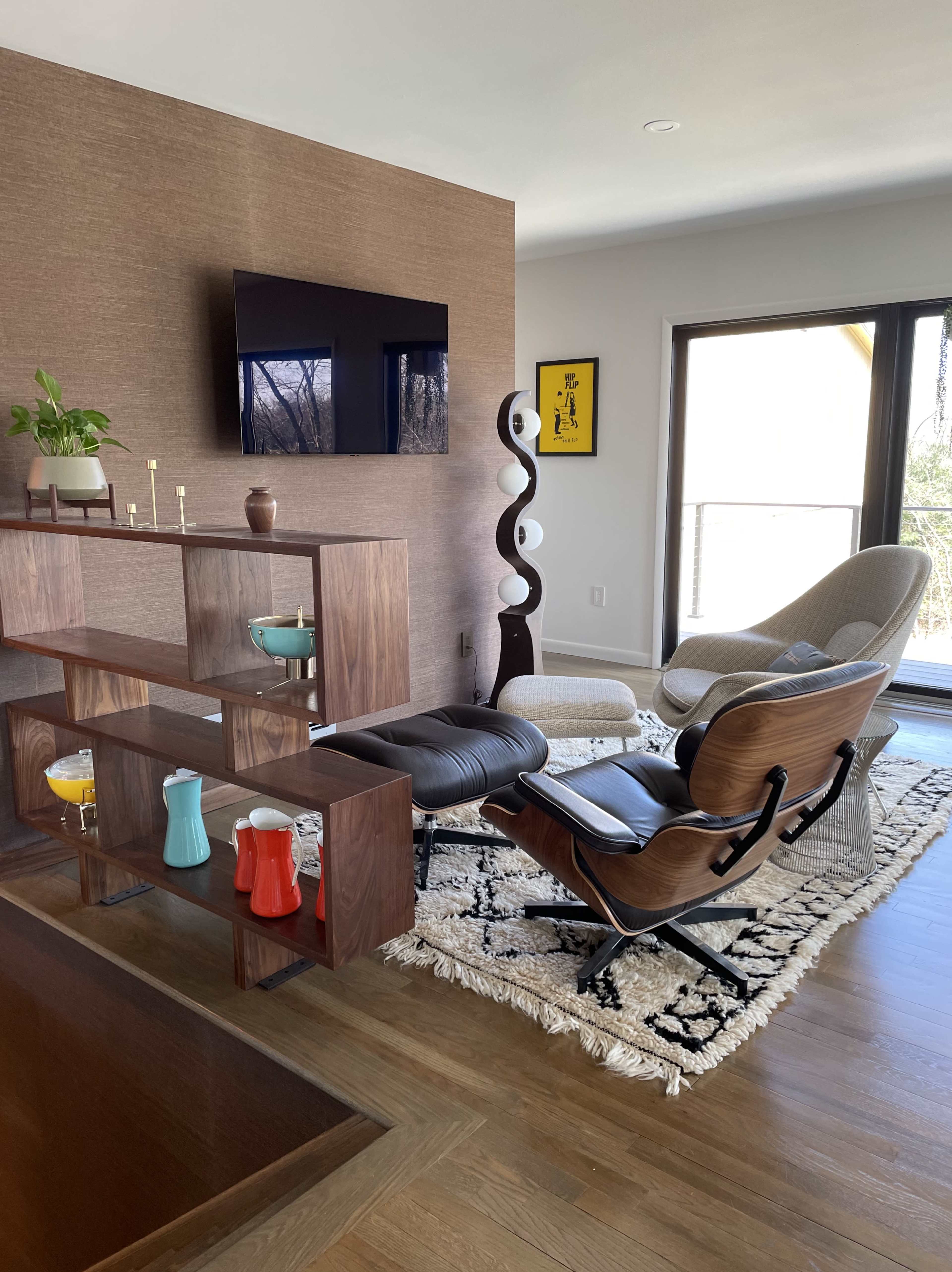 A modern living room with a wooden shelf displaying decorative items, a recliner chair, a television on the wall, and large windows allowing natural light to fill the space.