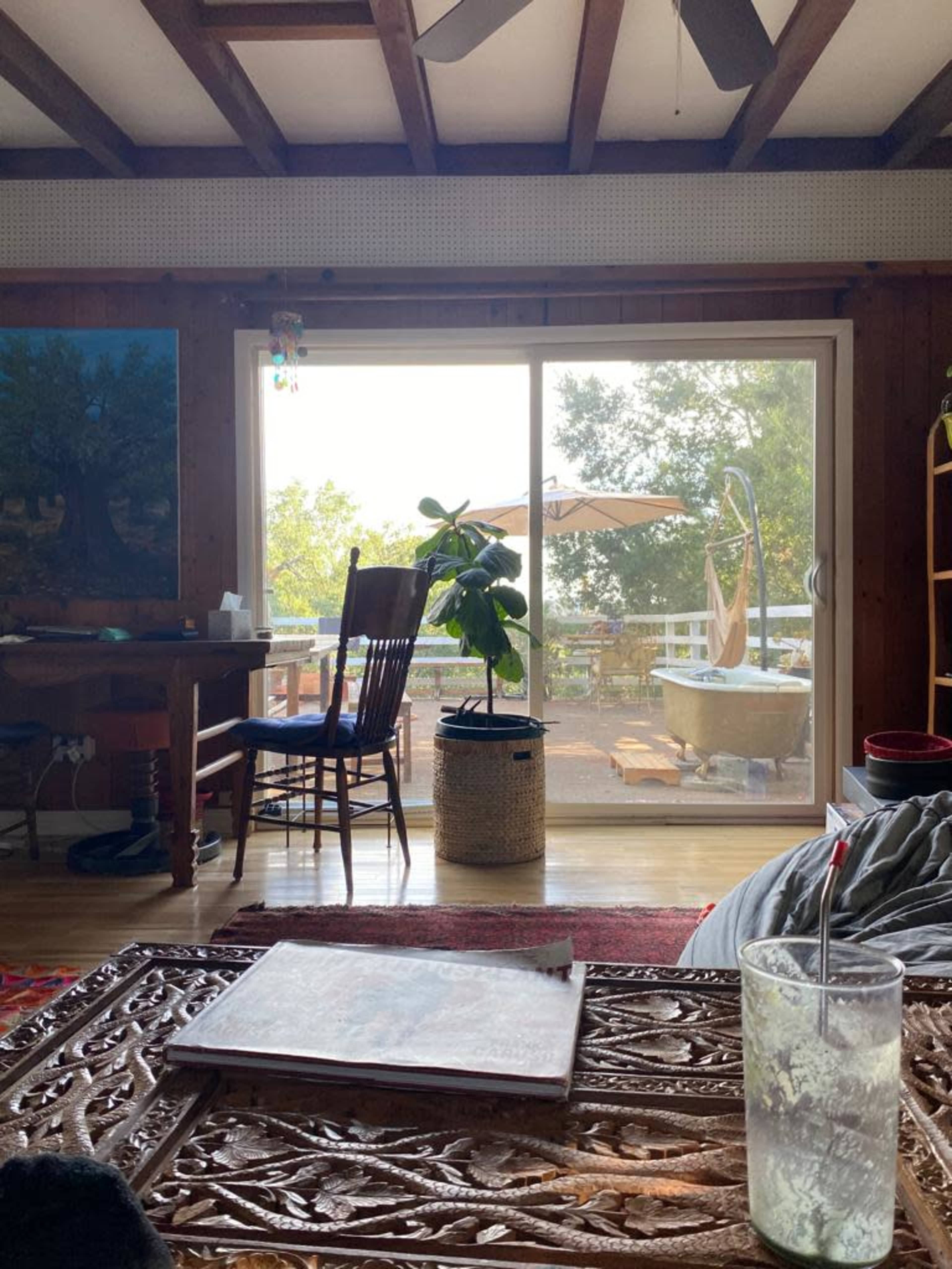 The image shows a cozy indoor space with wooden beams, a table and chair, a potted plant, and a large sliding glass door that leads to an outdoor area with a hot tub.
