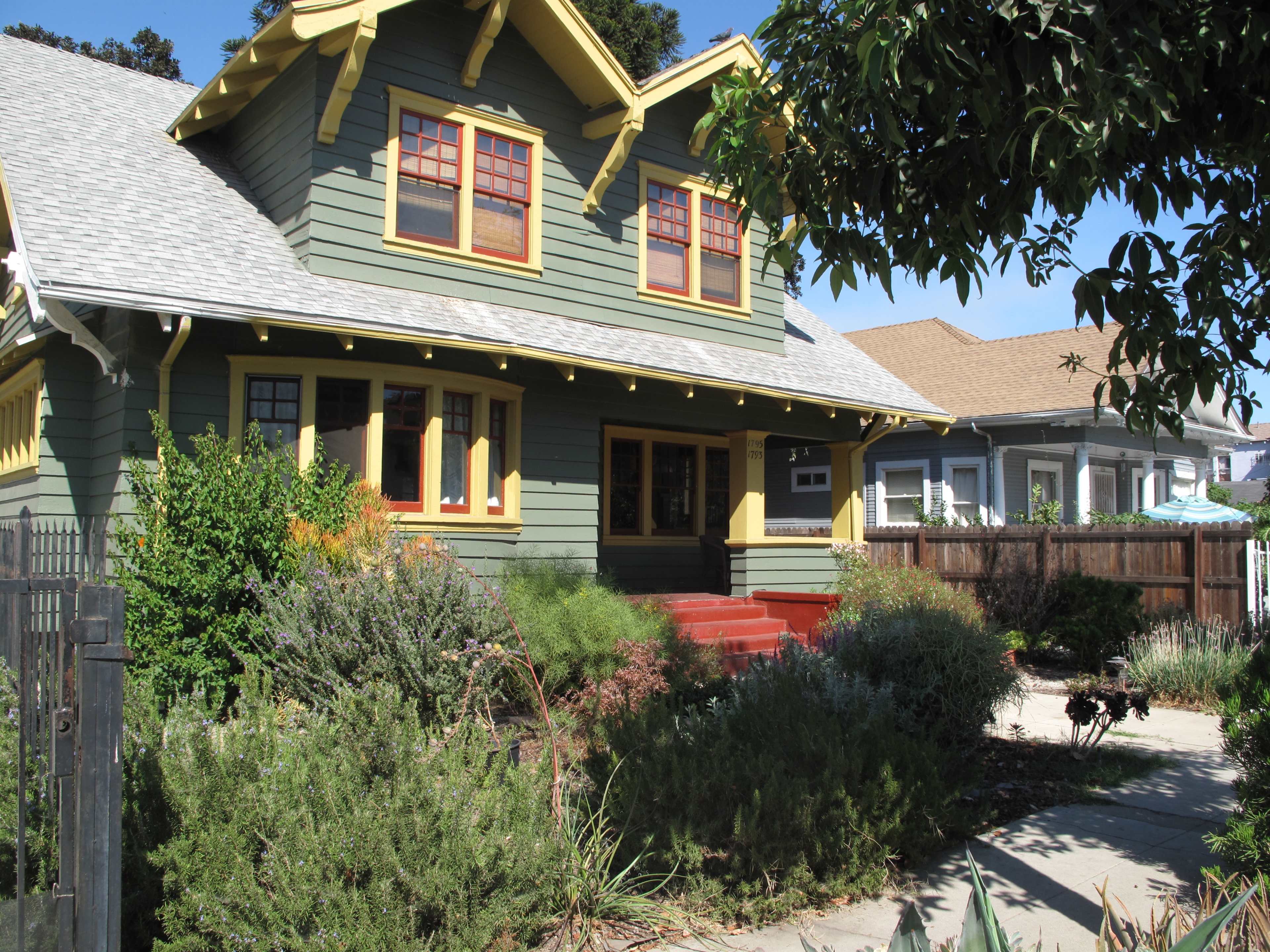 A two-story green house with yellow trim and a front porch is surrounded by various plants and shrubs.