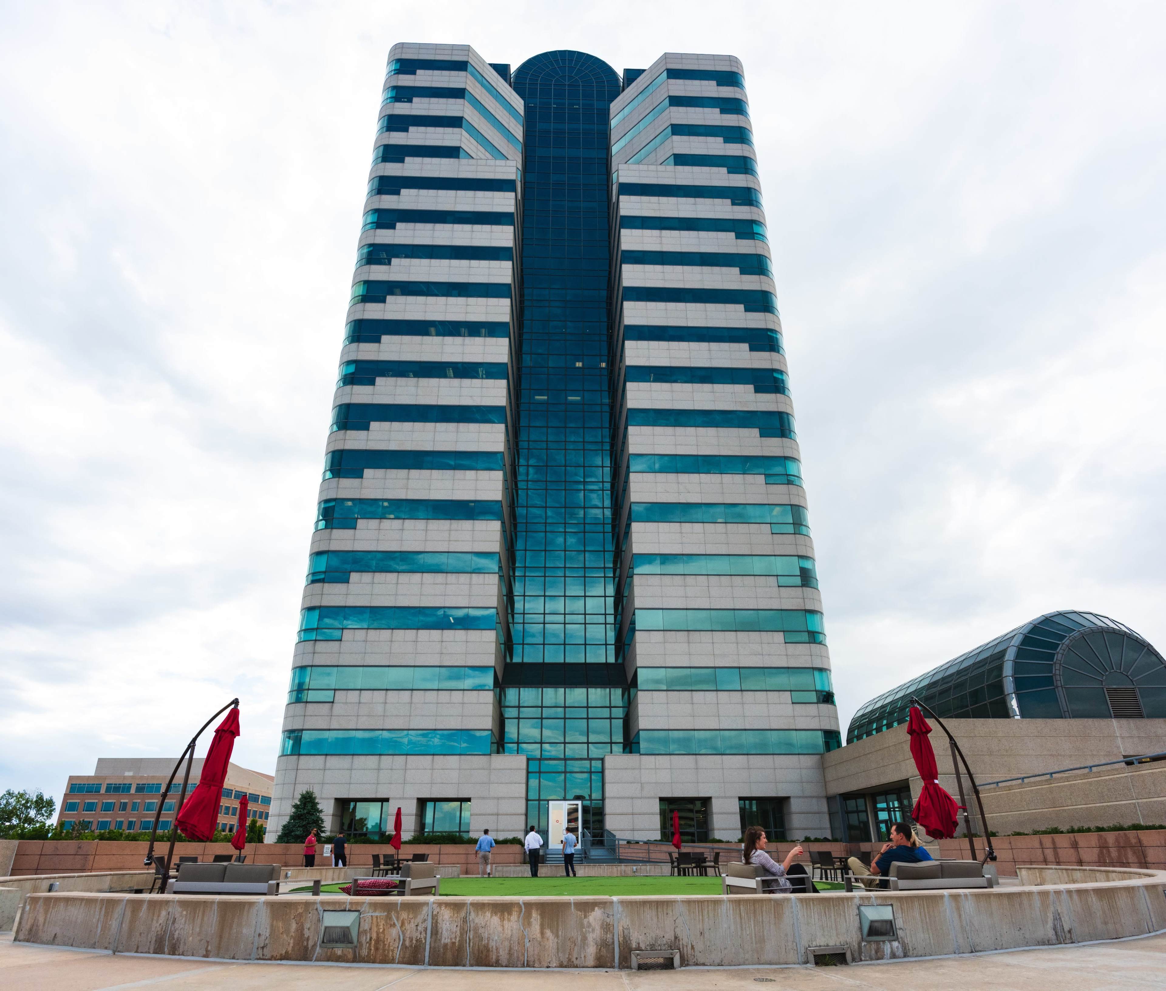A tall, modern office building with reflective glass windows is surrounded by a landscaped area featuring red umbrellas and seating.
