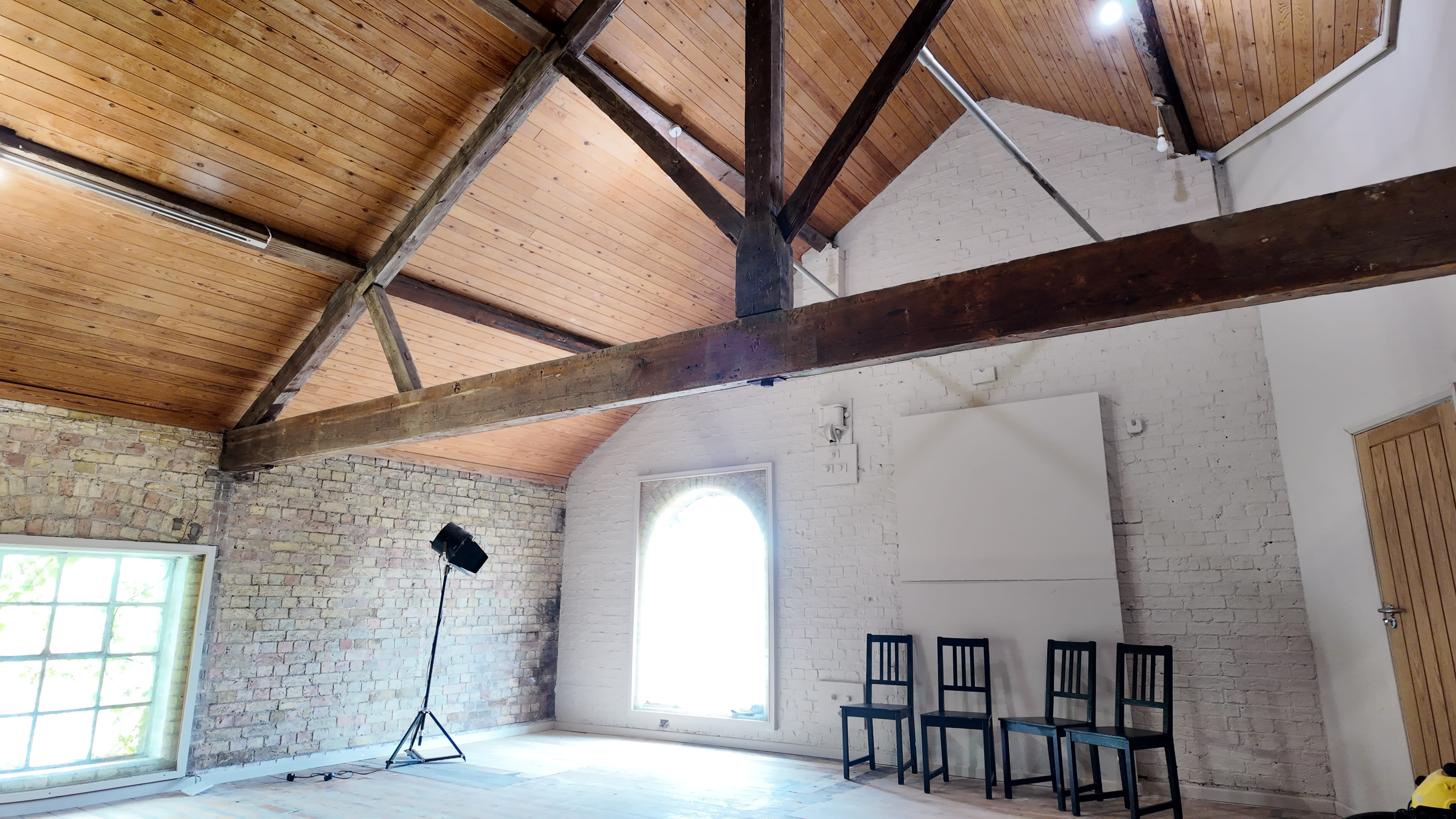 The image shows a spacious room with a high wooden ceiling, exposed beams, a large window, a blank wall, and three chairs arranged against it.