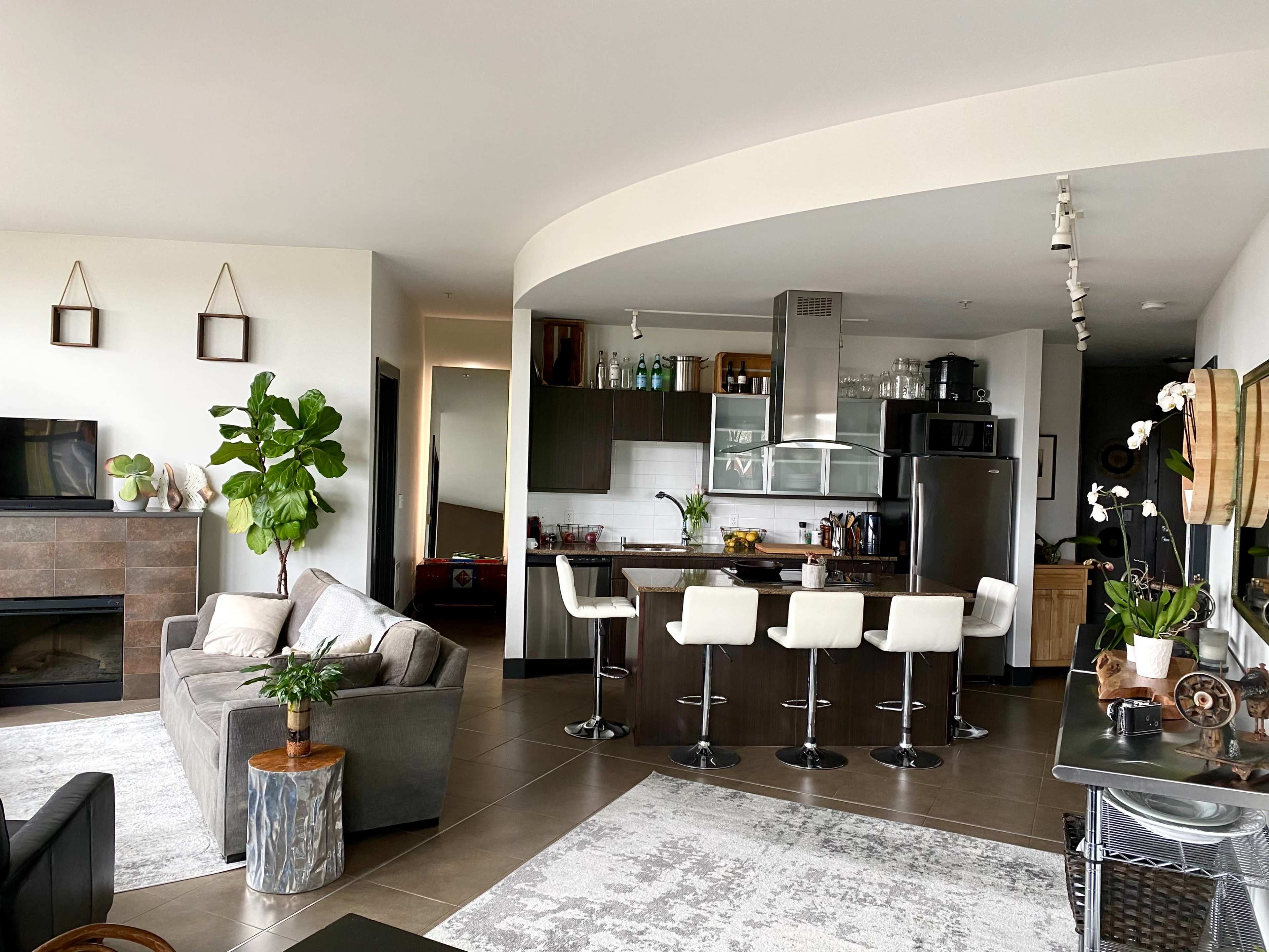 A modern open-concept living space featuring a kitchen bar with four stools, a fireplace, and plants, set against a backdrop of light-colored walls and large windows.