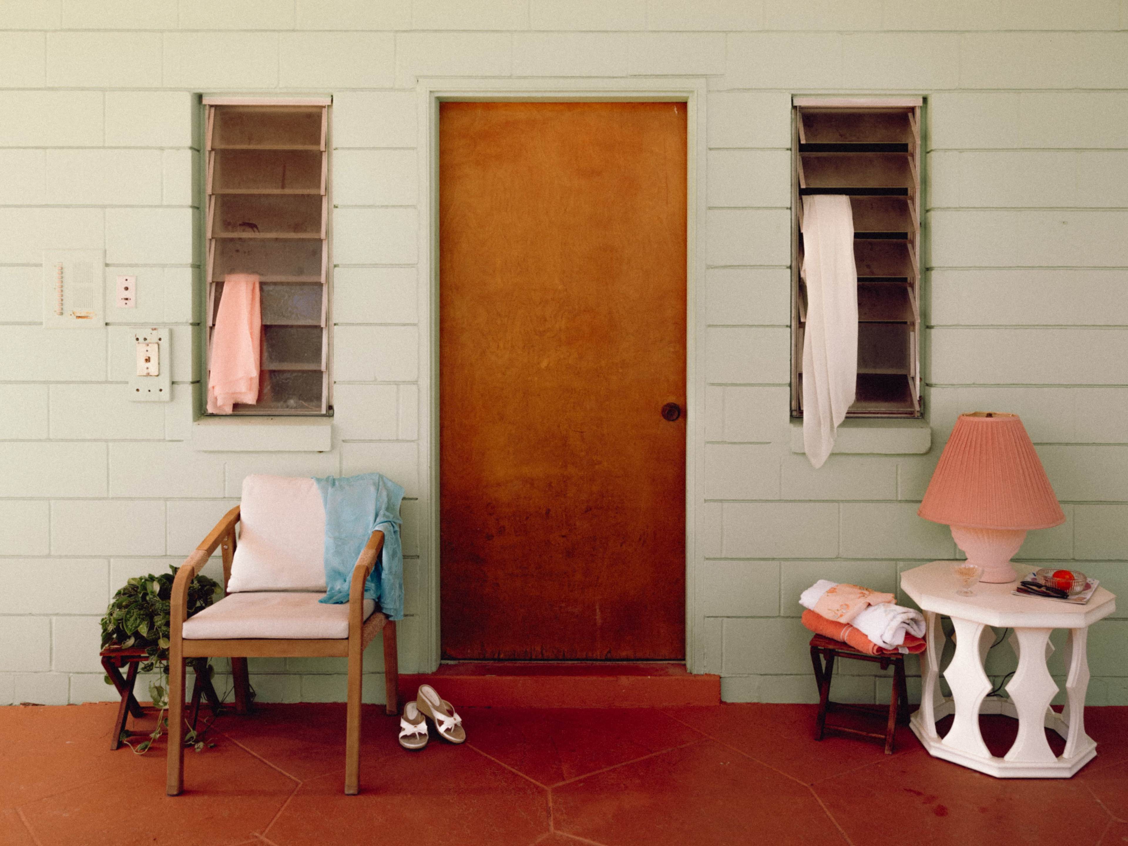 The image shows a wooden door flanked by two window units, with a chair, a small table with a lamp, and clothing items hanging nearby on a colored wall.