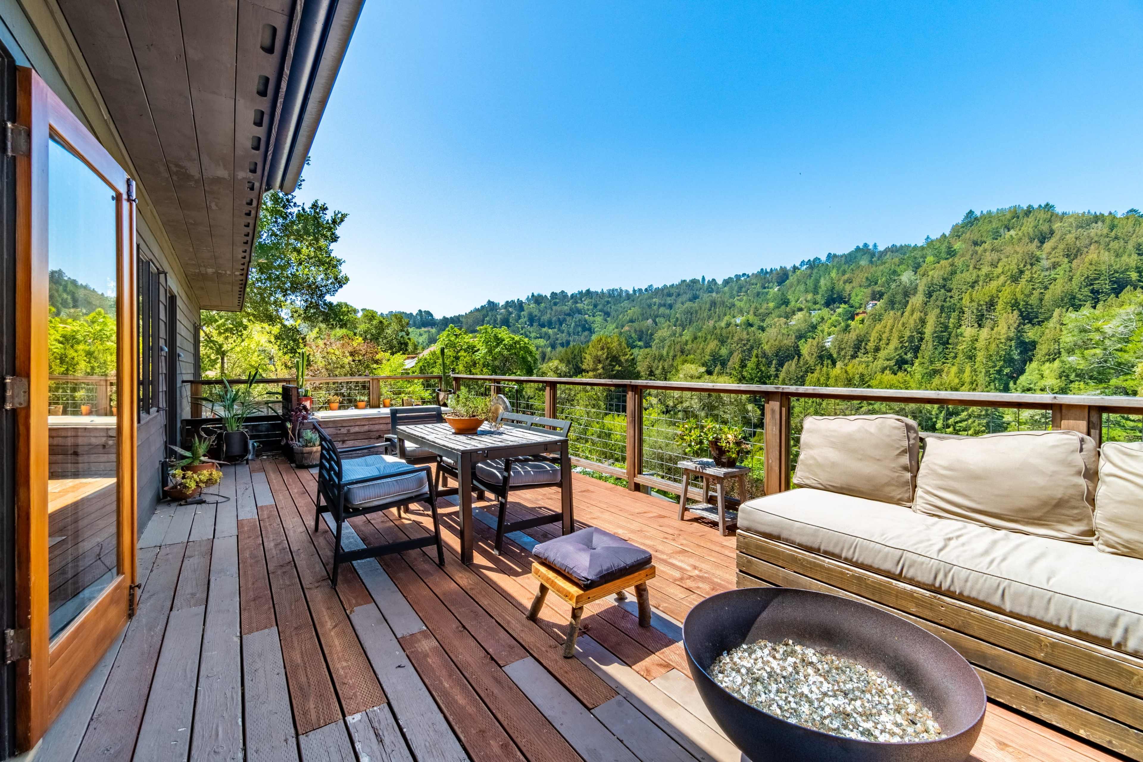 A wooden deck features a seating arrangement, table, and fire pit, overlooking a lush green hillside under a clear blue sky.