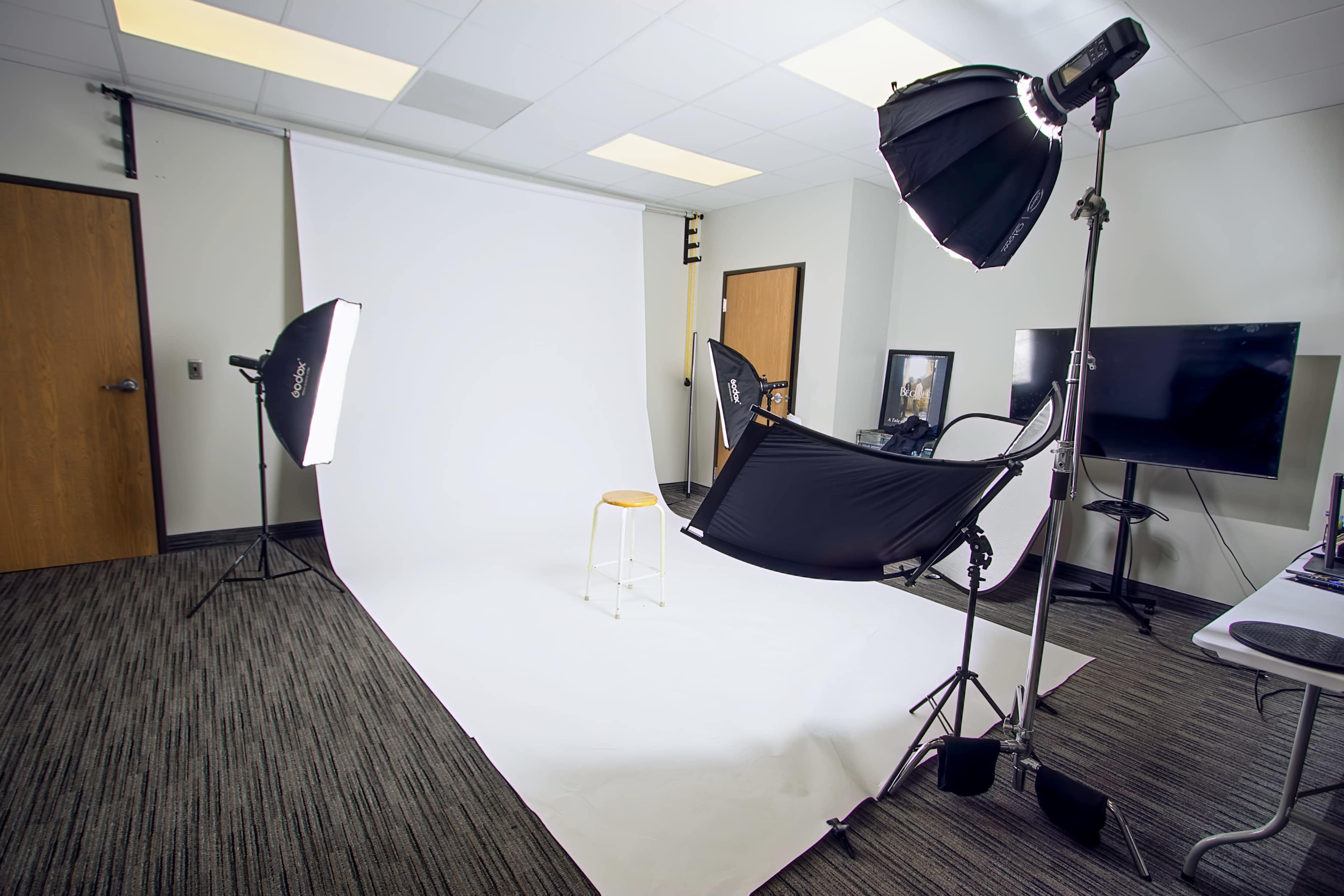 The image shows a photography studio with a white backdrop, a stool positioned in front, and several lighting equipment setups.