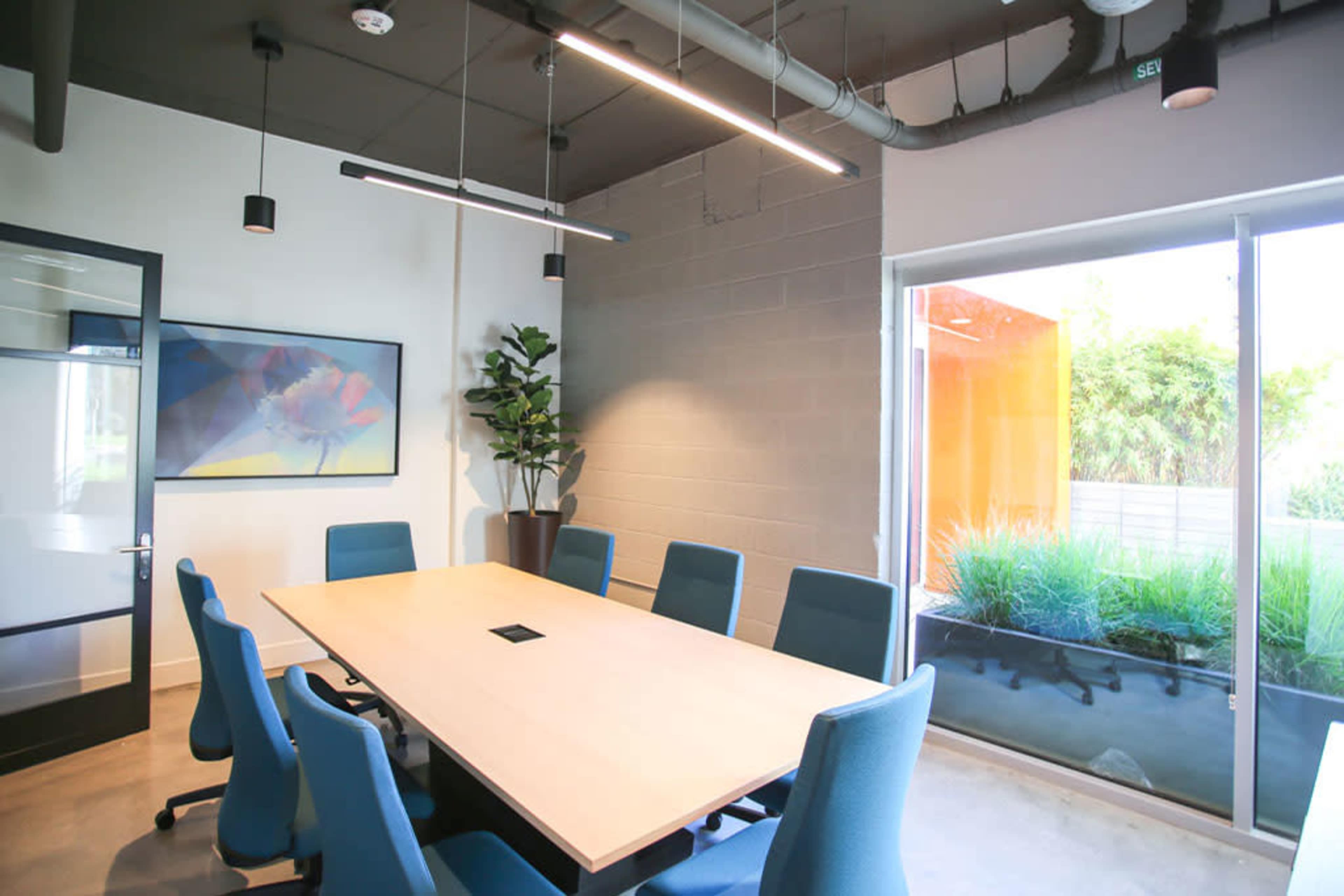 A modern conference room features a large wooden table surrounded by blue chairs, with a plant and a glass wall showing a view of greenery outside.