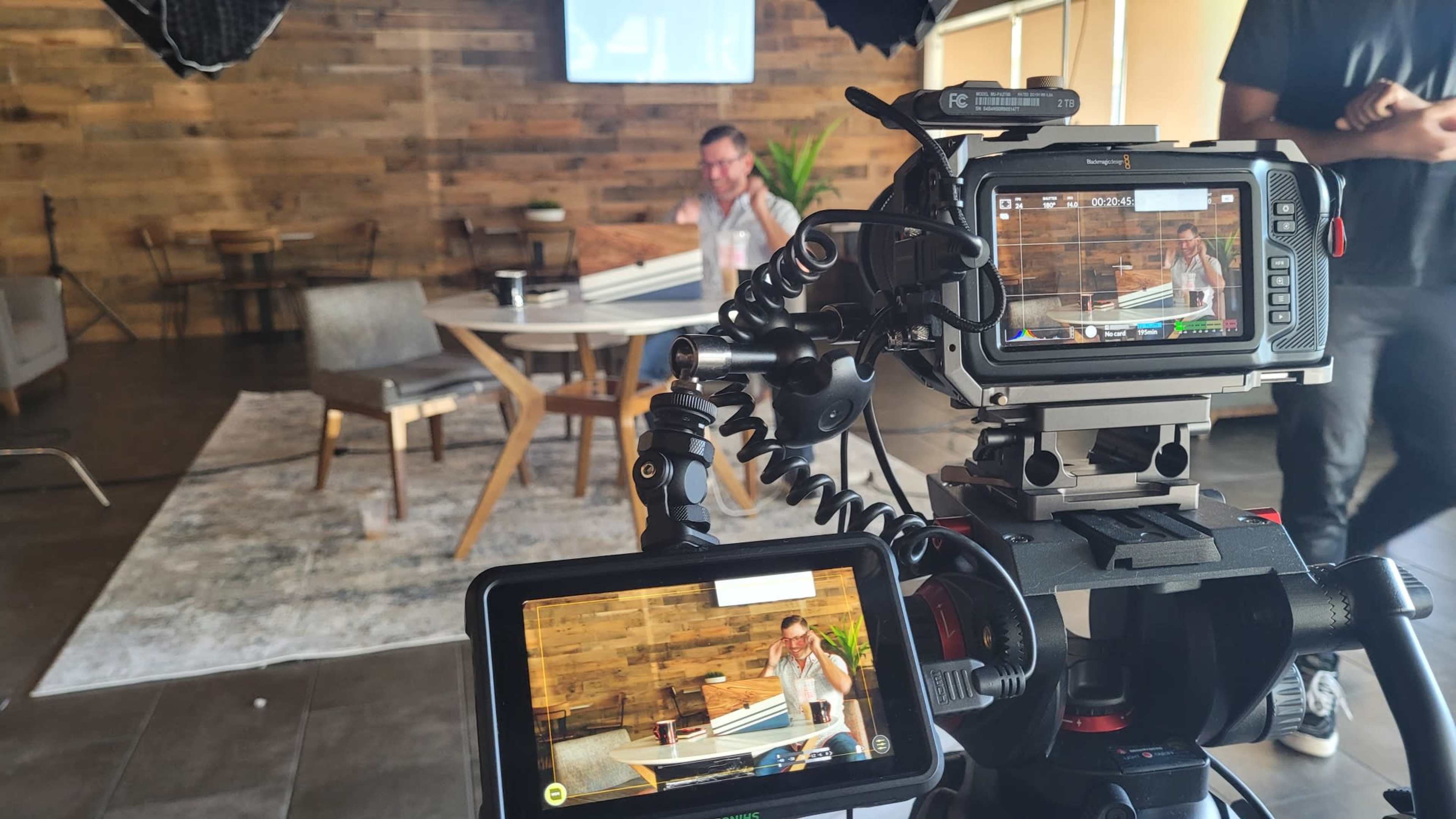 A camera setup captures a person seated at a table in a room with wooden walls and modern furniture.