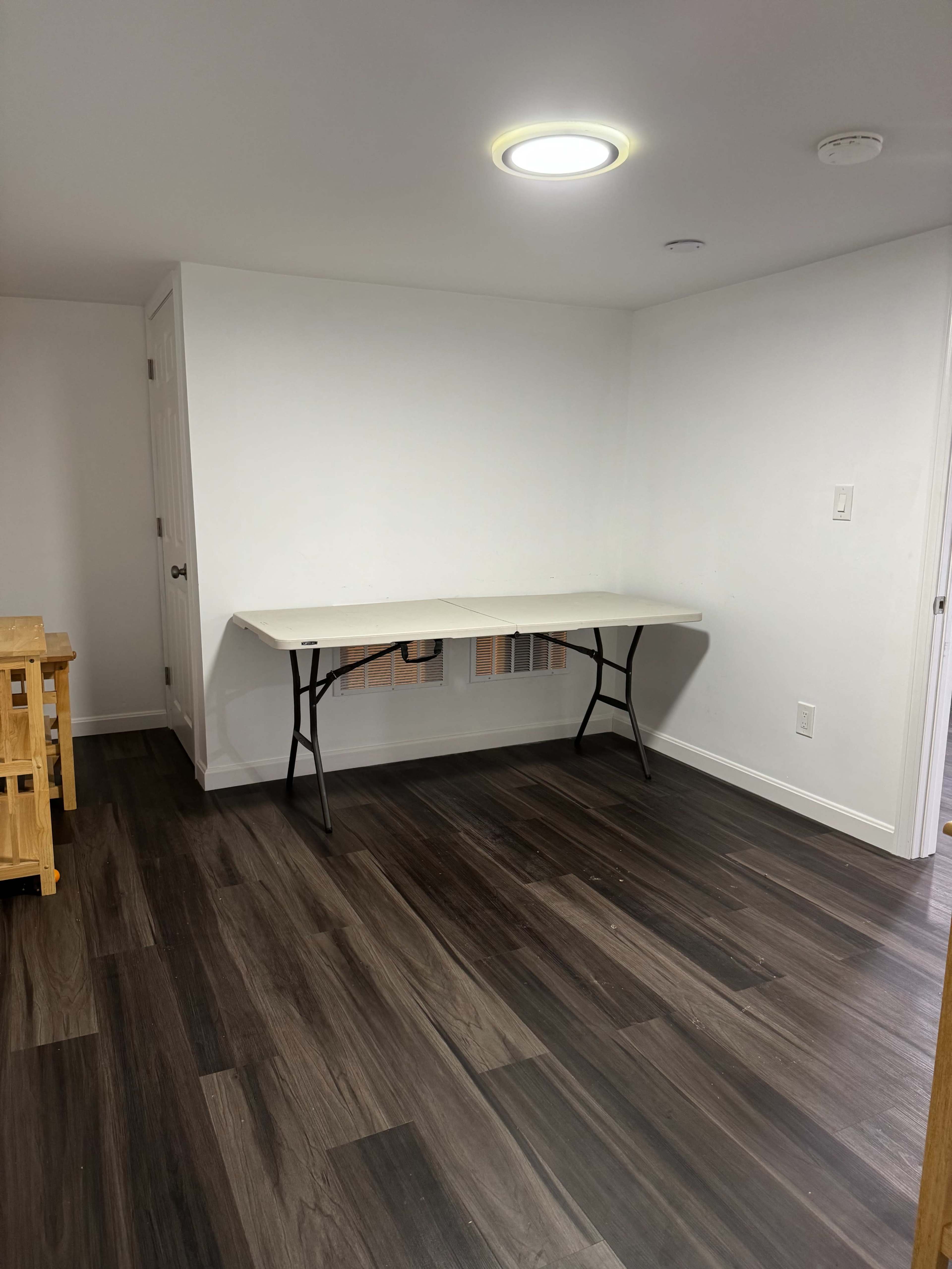 The image shows a sparsely furnished room with a foldable table set up against a white wall and a door in the background.