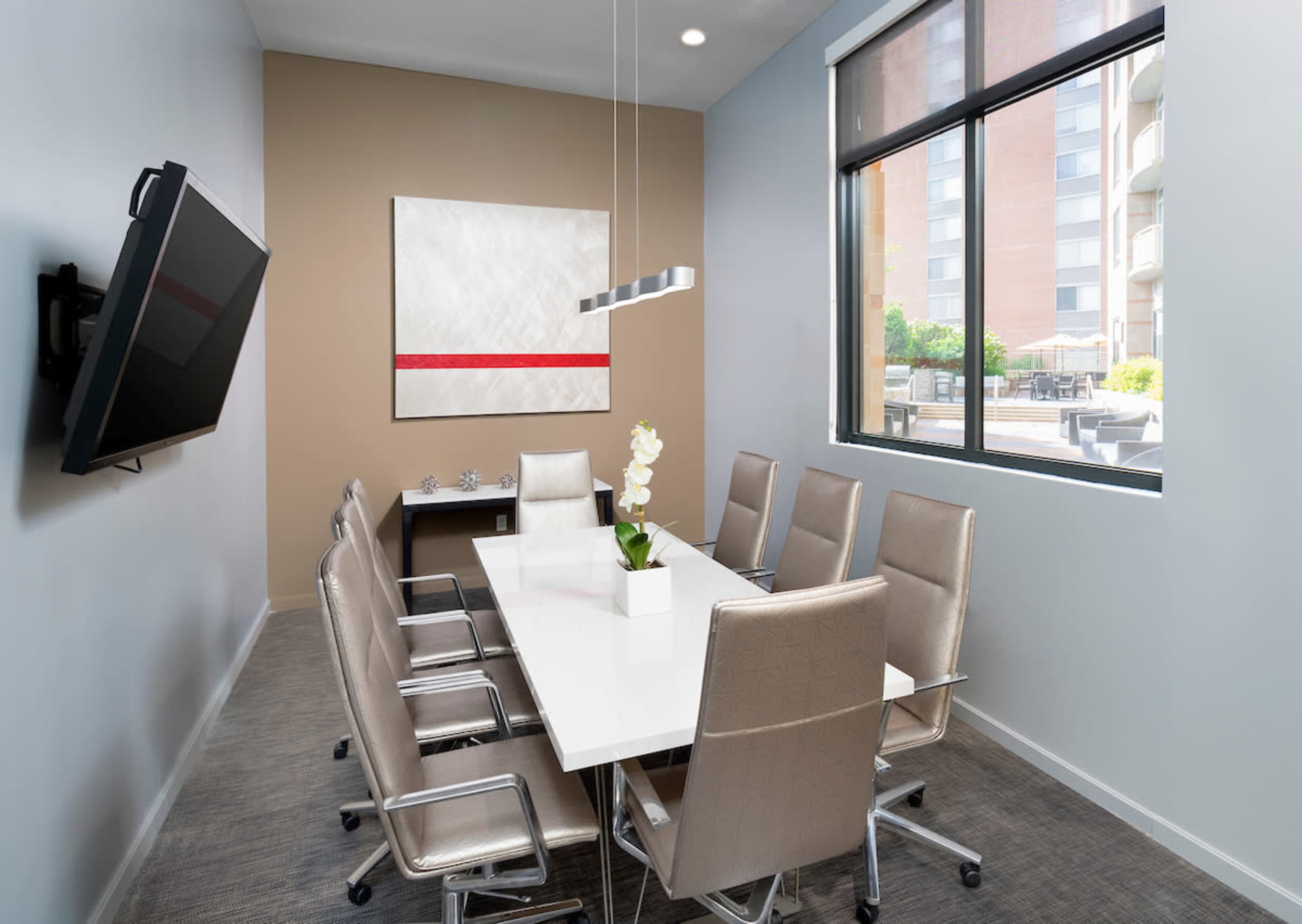 A modern conference room features a long white table surrounded by six silver chairs, with a television mounted on the wall and a large window providing natural light.