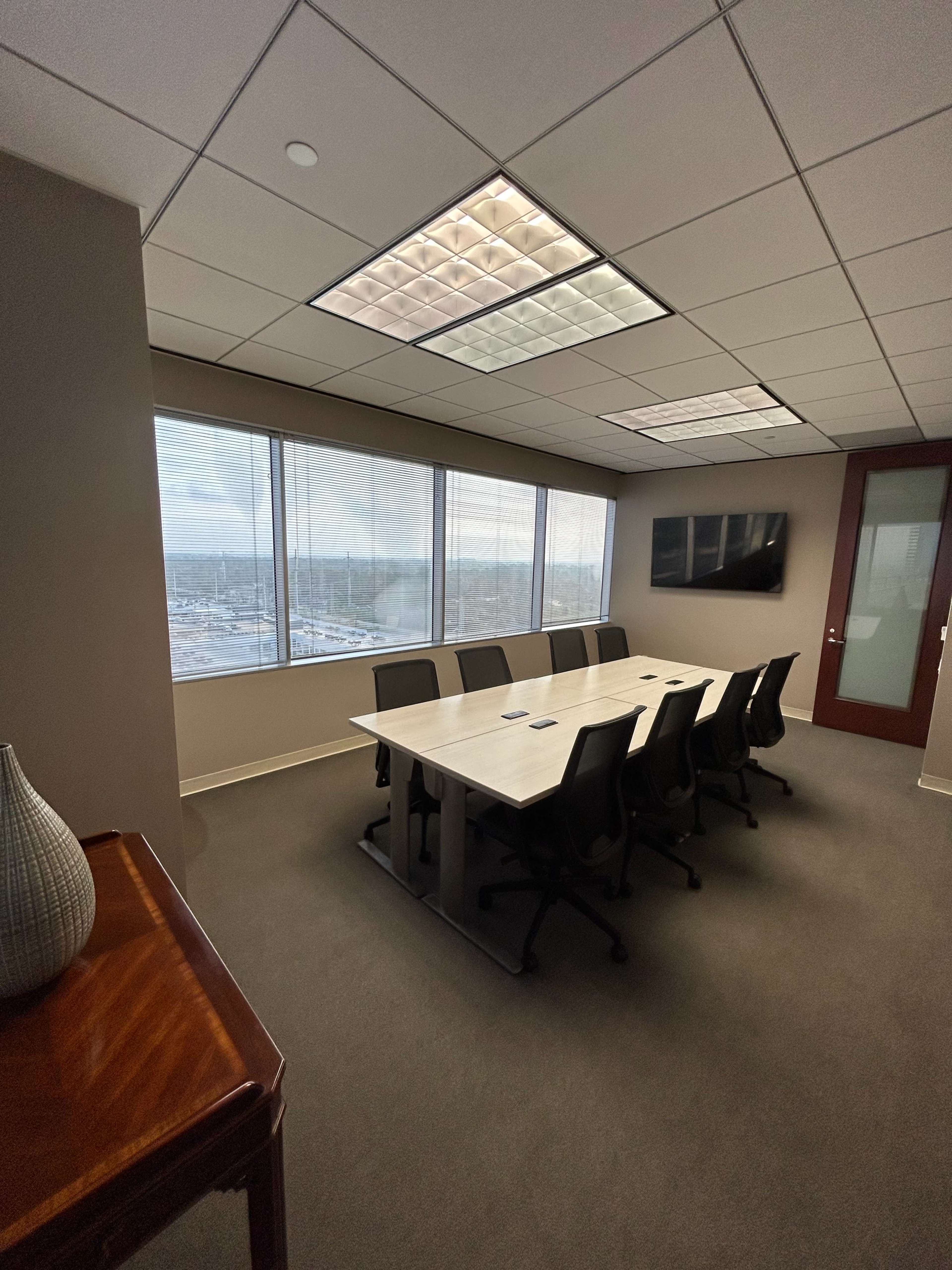 The image shows a modern conference room with a long table, eight chairs, a large window with a view, and a wall-mounted screen.