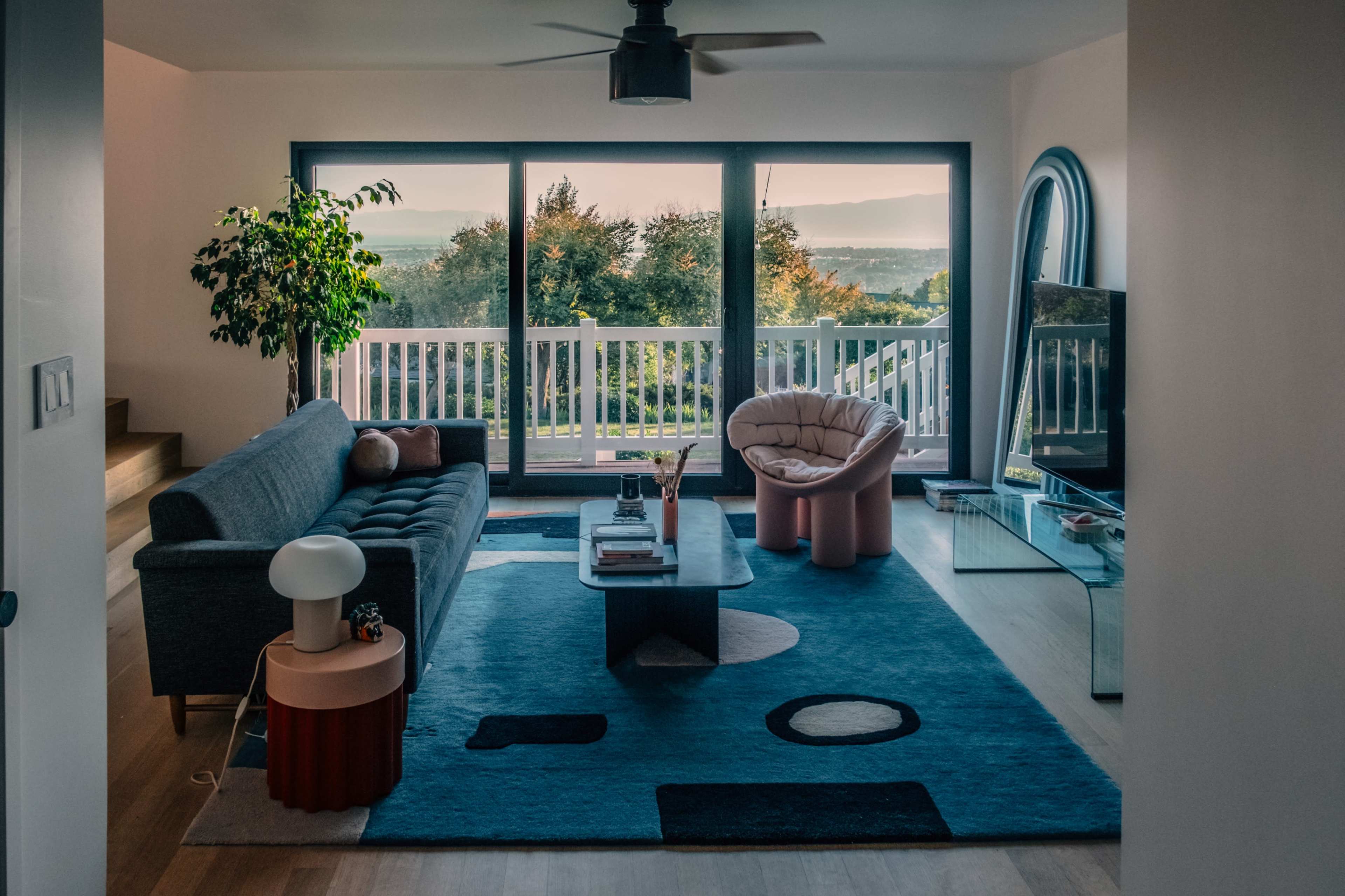 The image shows a modern living room with a large window revealing a view of greenery, a gray sofa, a pink armchair, and a coffee table on a colorful rug.