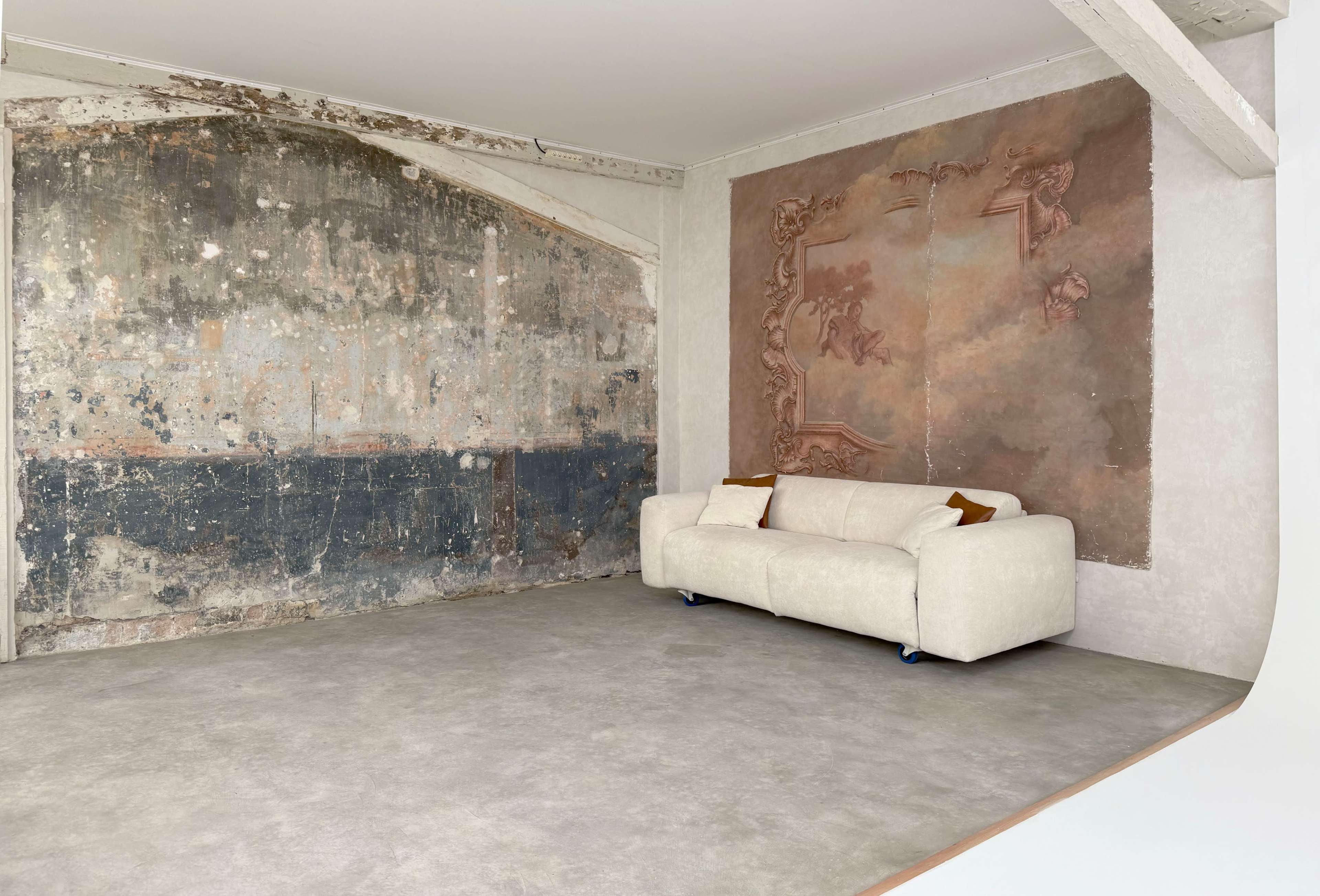 The image shows a minimalist interior with a worn, textured wall on one side and a modern white sofa placed on a smooth floor.