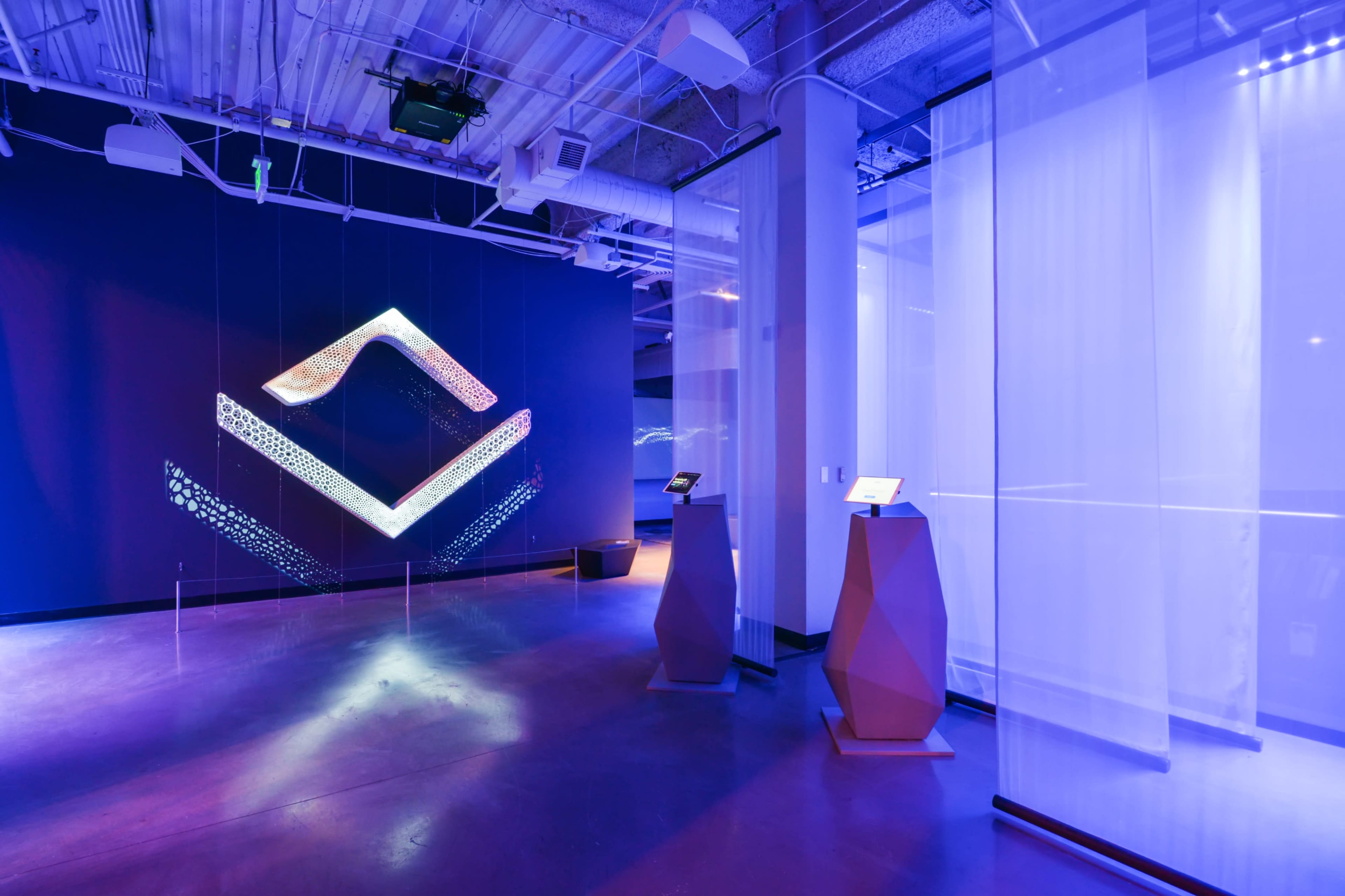 An art installation in a modern exhibition space featuring geometric light projections on a blue wall and two stylized pedestals displaying interactive screens.