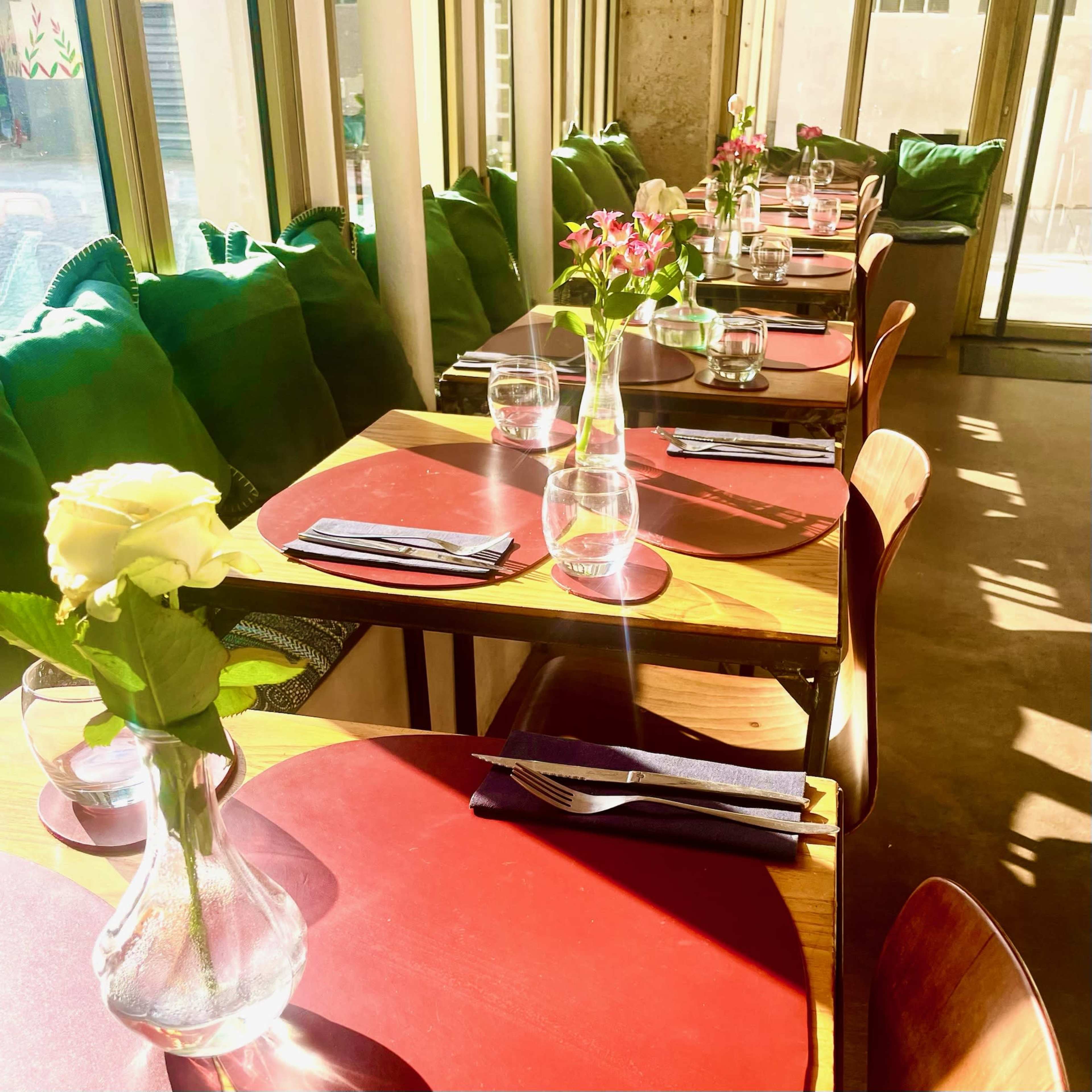 The image shows a neatly arranged dining area with tables set for guests, featuring vases of flowers, water glasses, and green cushions along the windows.