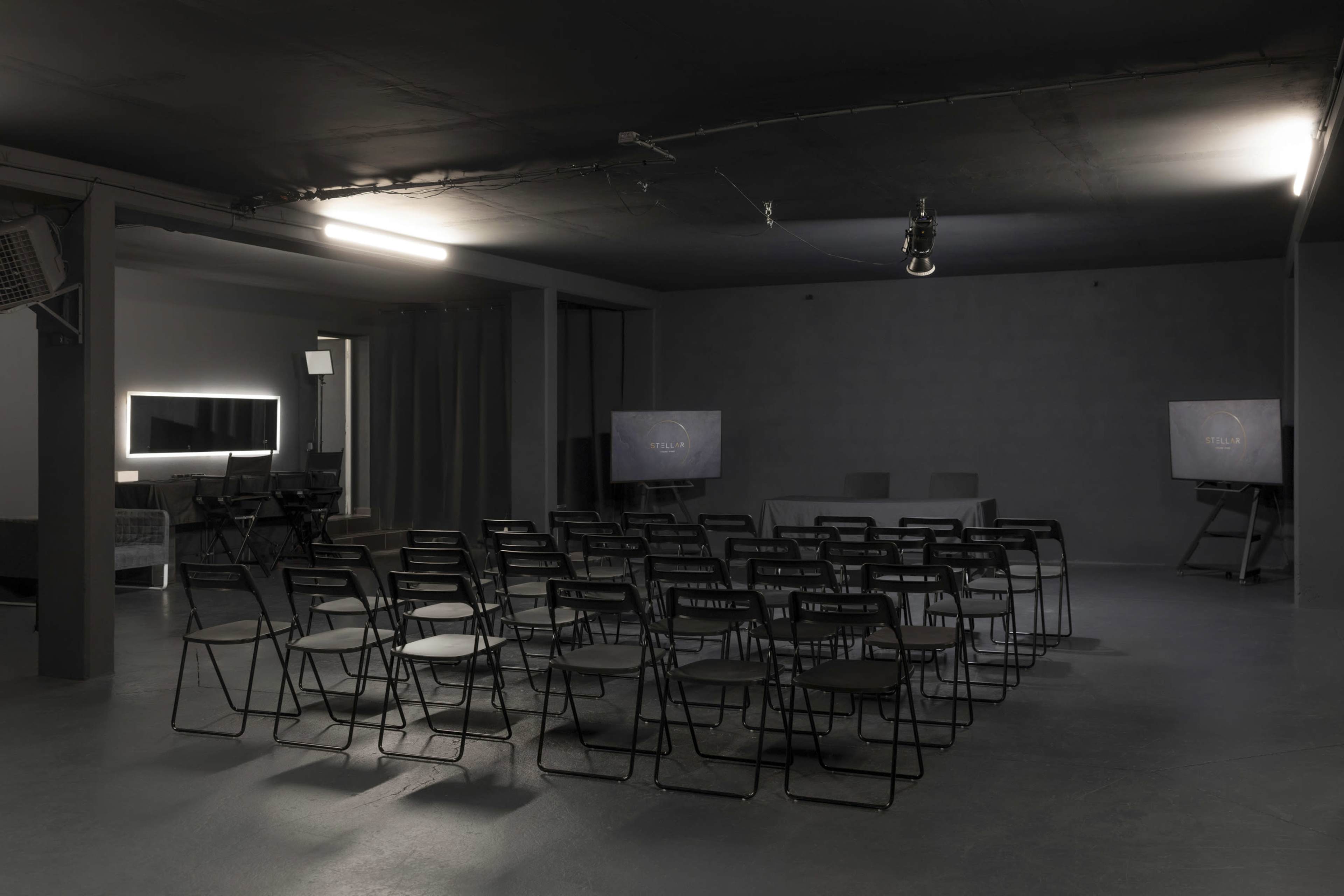 A dimly lit room features rows of black chairs facing two screens, with a table set in the background.
