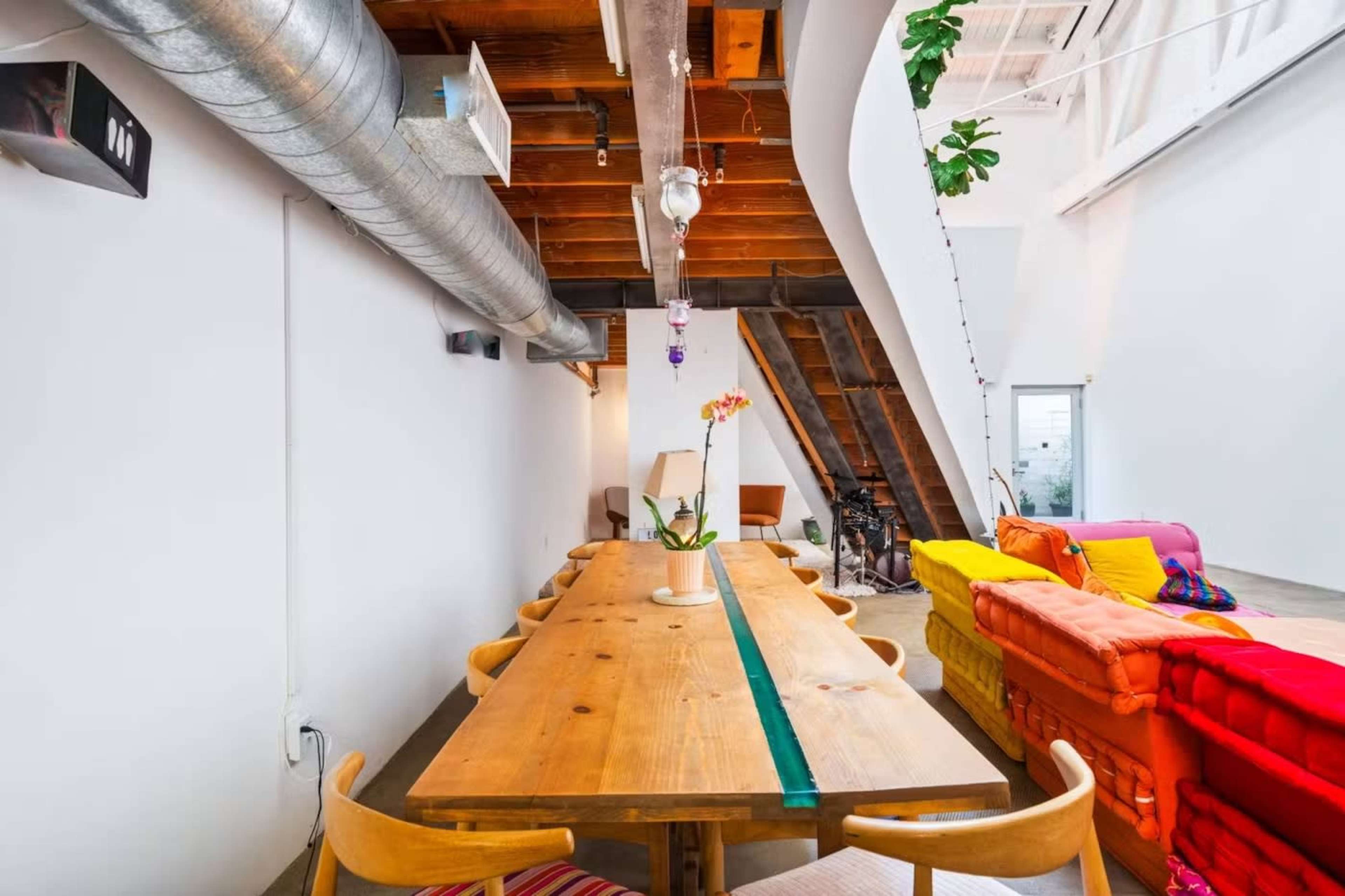 A long wooden dining table is set in a room with exposed wooden beams, colorful sofas, and a potted plant.