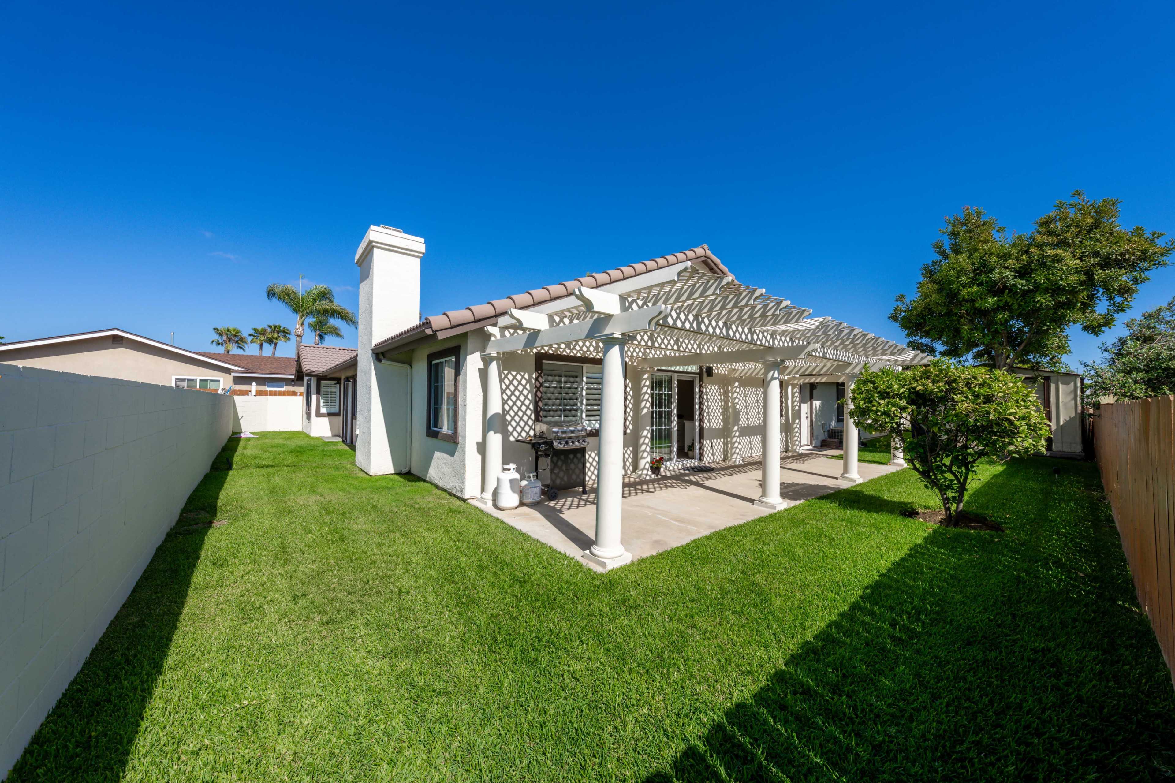 A single-story house features a covered patio with a pergola, surrounded by green grass and landscaping.