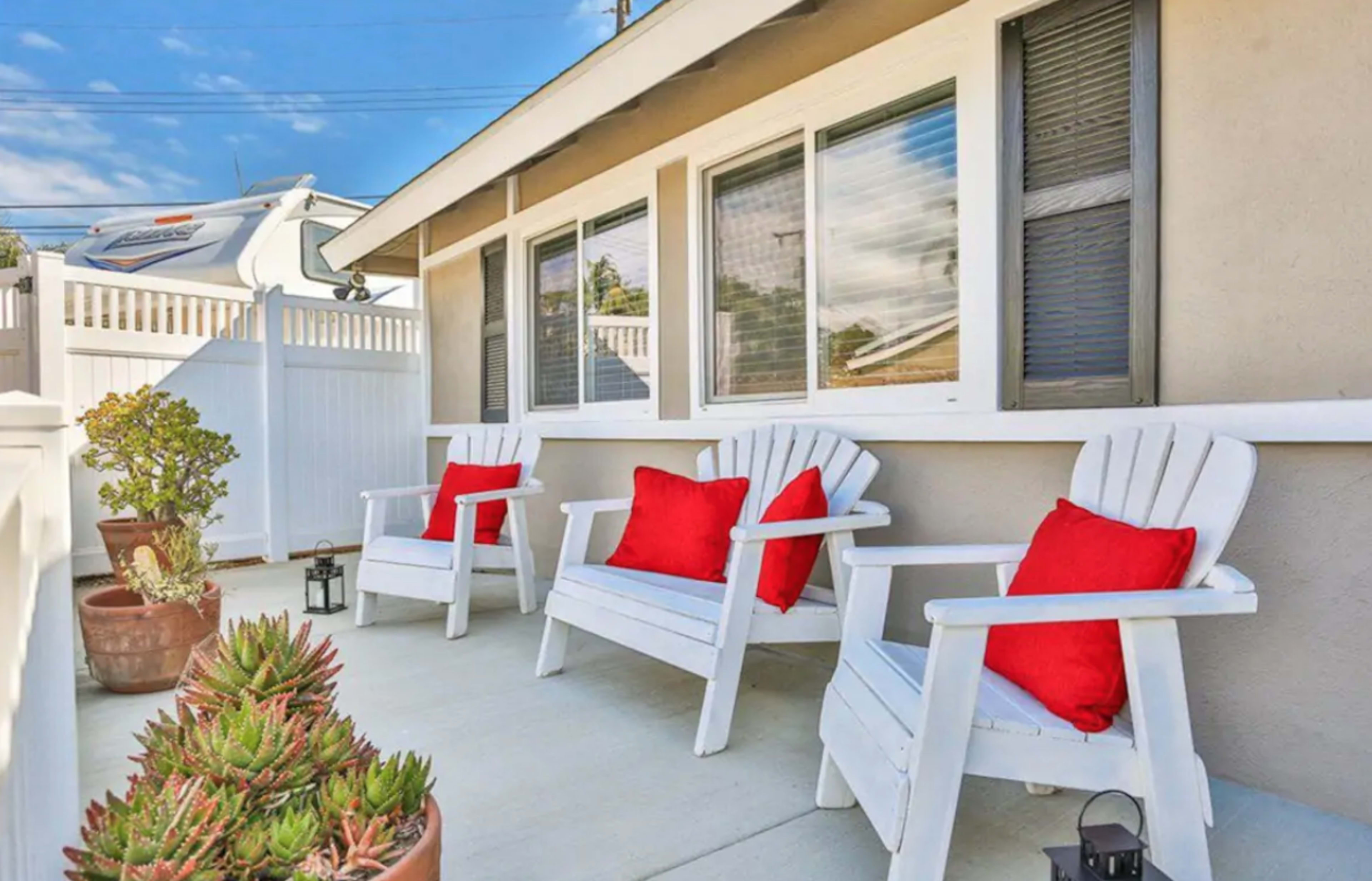 A patio area with three white wooden chairs, each adorned with a red cushion, surrounded by potted plants and a decorative fence.