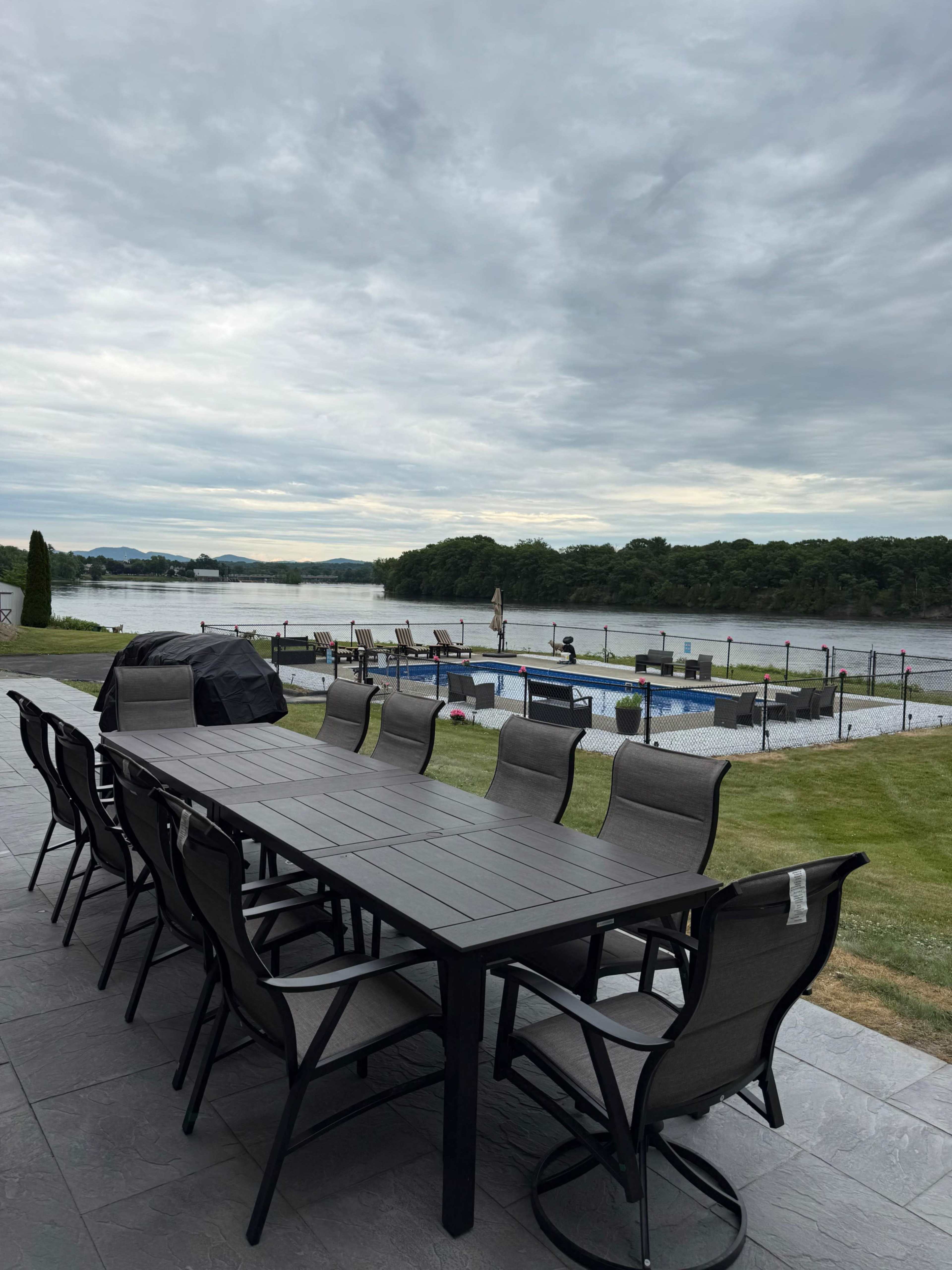 A large dining table with chairs is set up on a patio overlooking a pool and a river under a cloudy sky.