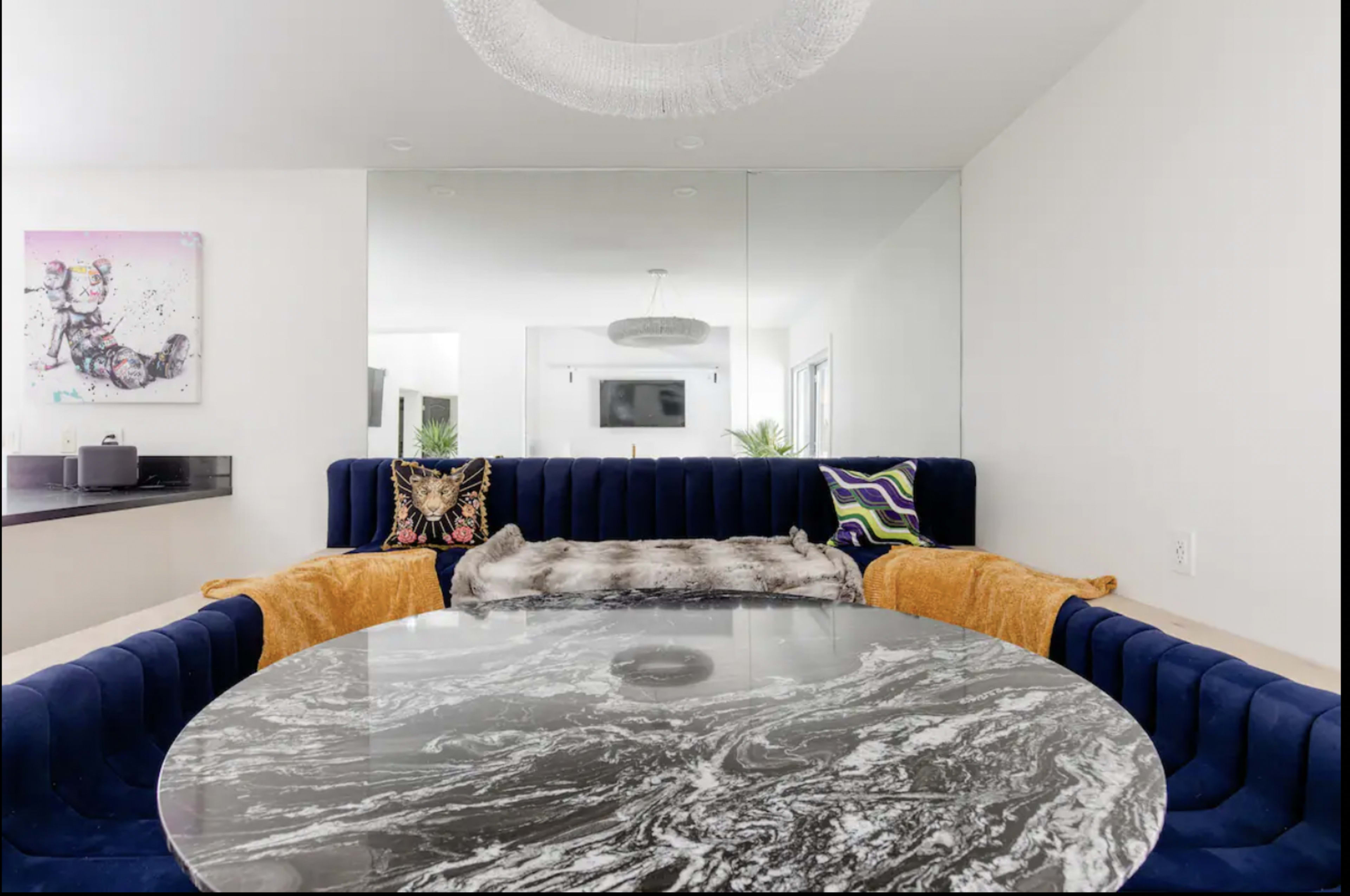 A dining area features a round marble table surrounded by a blue velvet sectional sofa with decorative pillows and a plush throw.