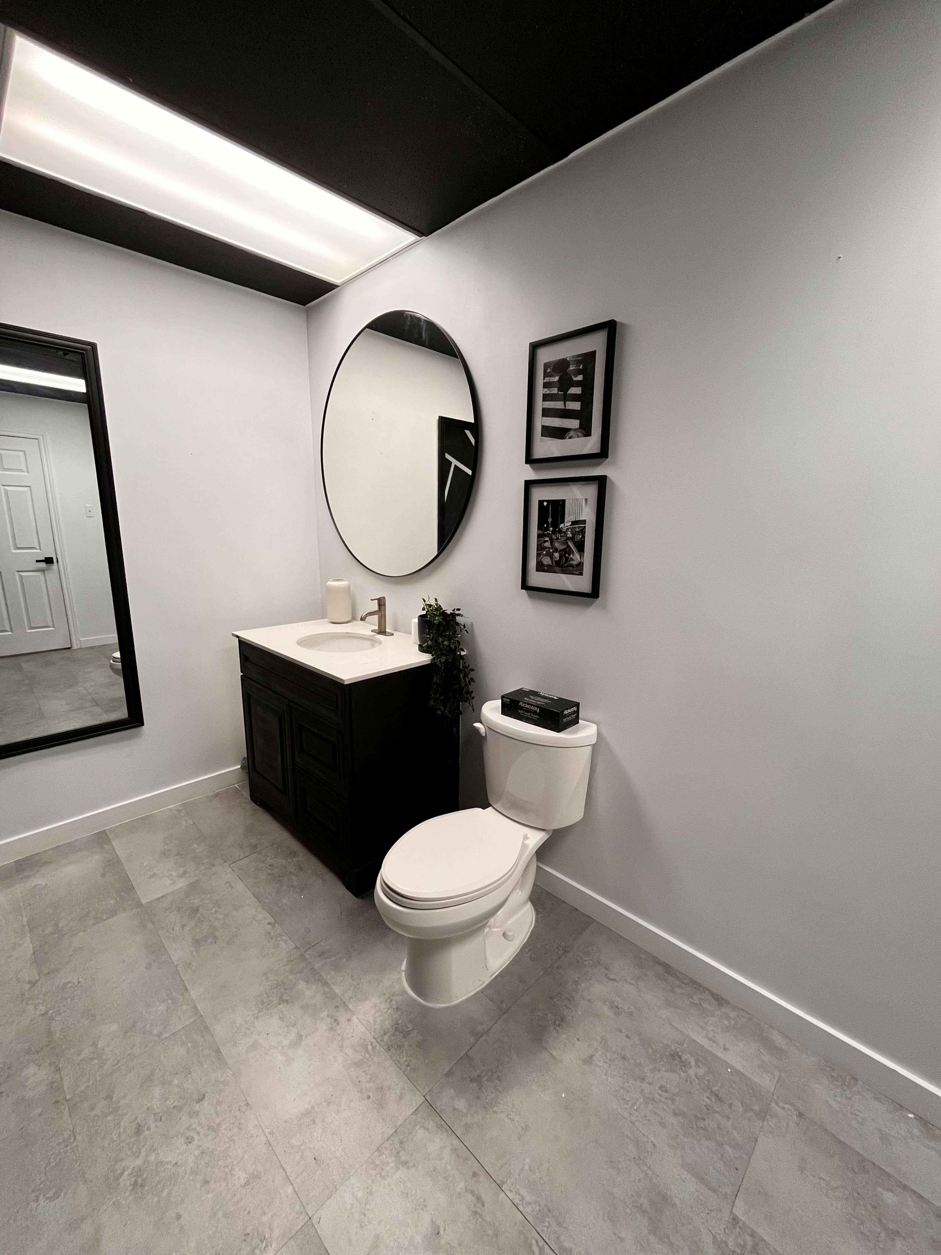 A modern bathroom with a round mirror above a dark vanity, a toilet, and framed wall art, set against light-colored walls and tiled flooring.