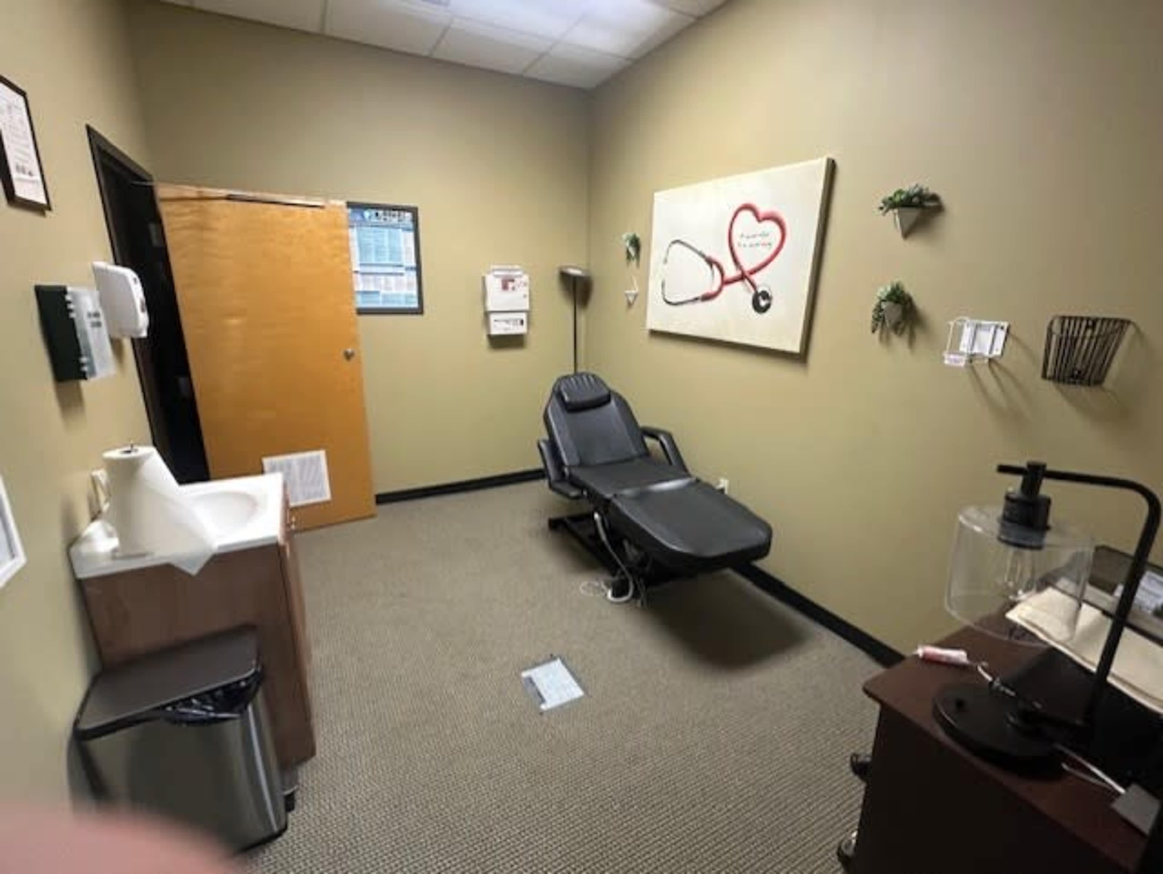 The image shows a small medical examination room with a reclining chair, a sink, a mini-fridge, and a wall featuring a framed heart and stethoscope artwork.