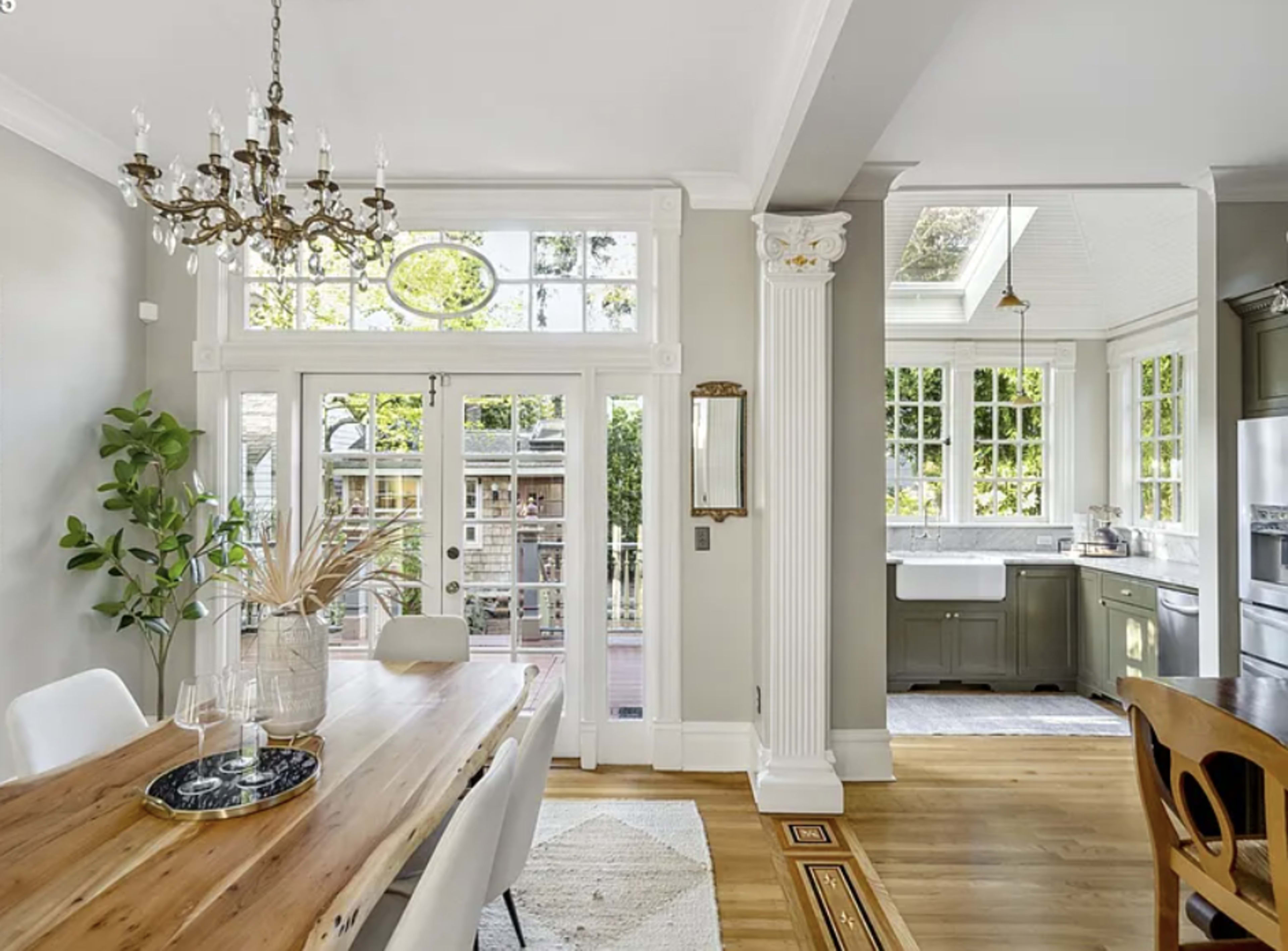 The image shows a bright, contemporary dining area with a wooden table, a chandelier, and large windows that overlook a backyard, while the kitchen is visible in the background.