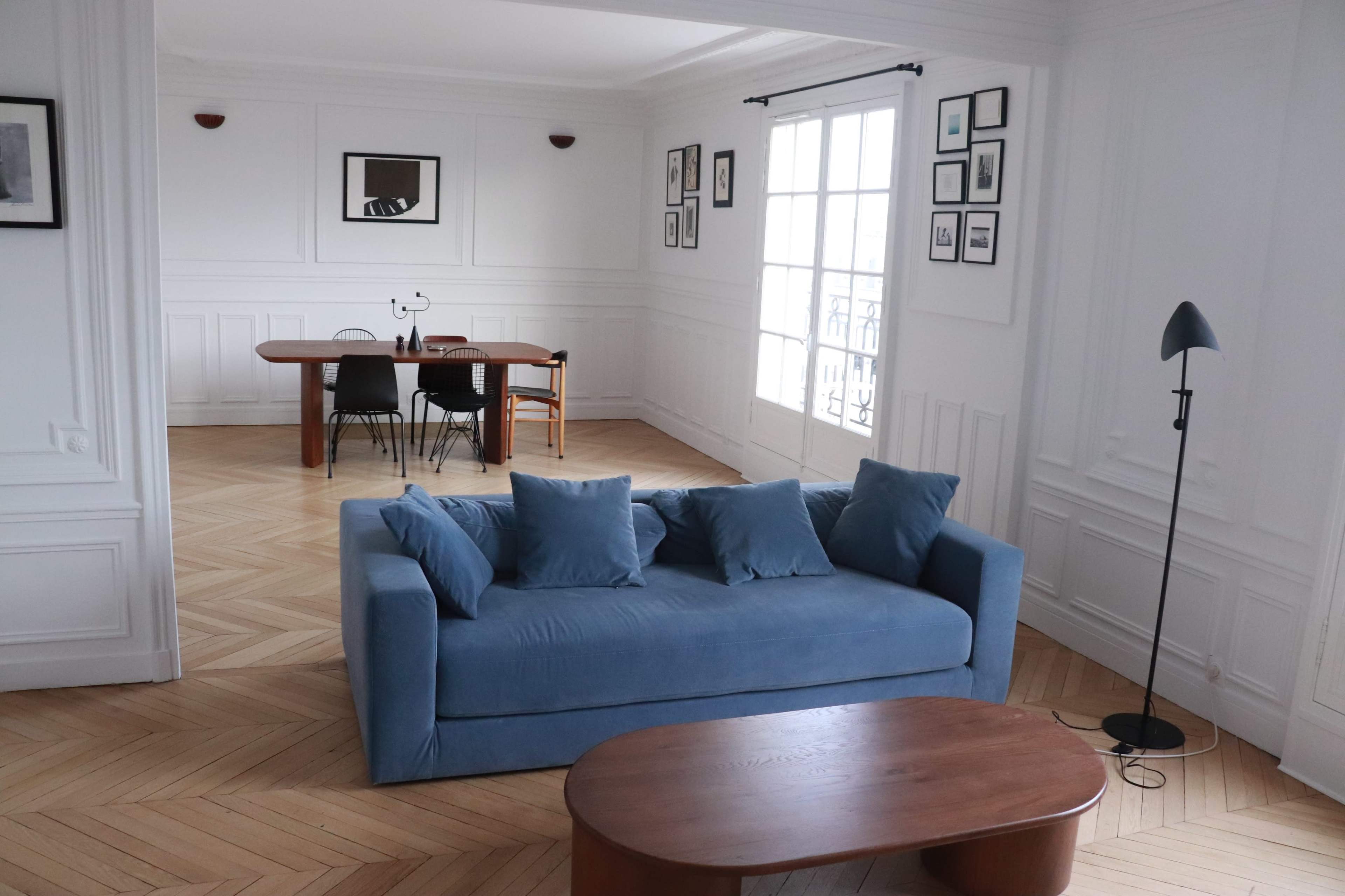 The image shows a spacious, well-lit room featuring a blue sofa, a wooden coffee table, and a dining area with a table and chairs against a wall adorned with framed pictures.