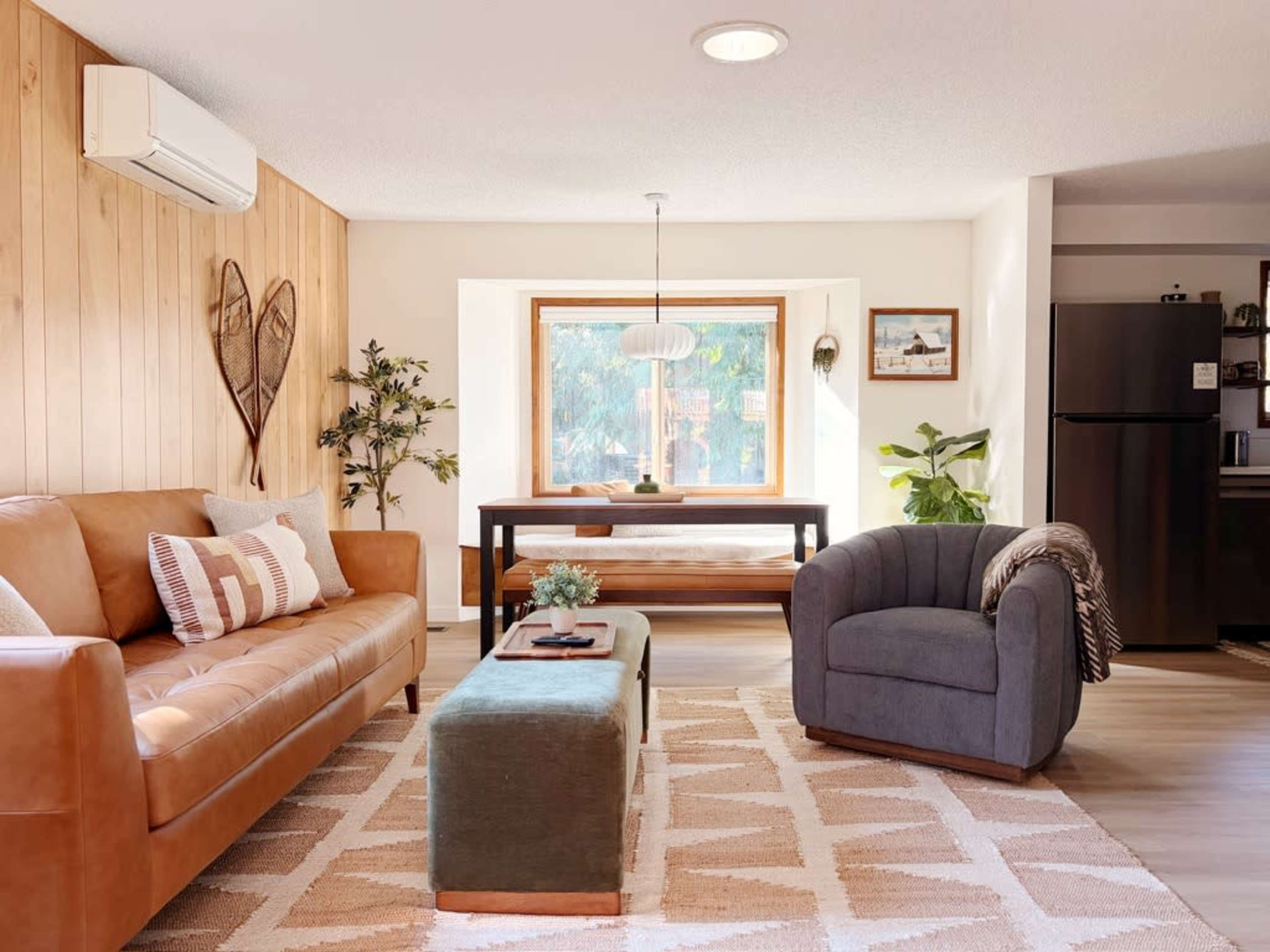 The scene features a cozy living room with a brown sofa, a round gray armchair, a wooden dining table, and a window that lets in natural light.