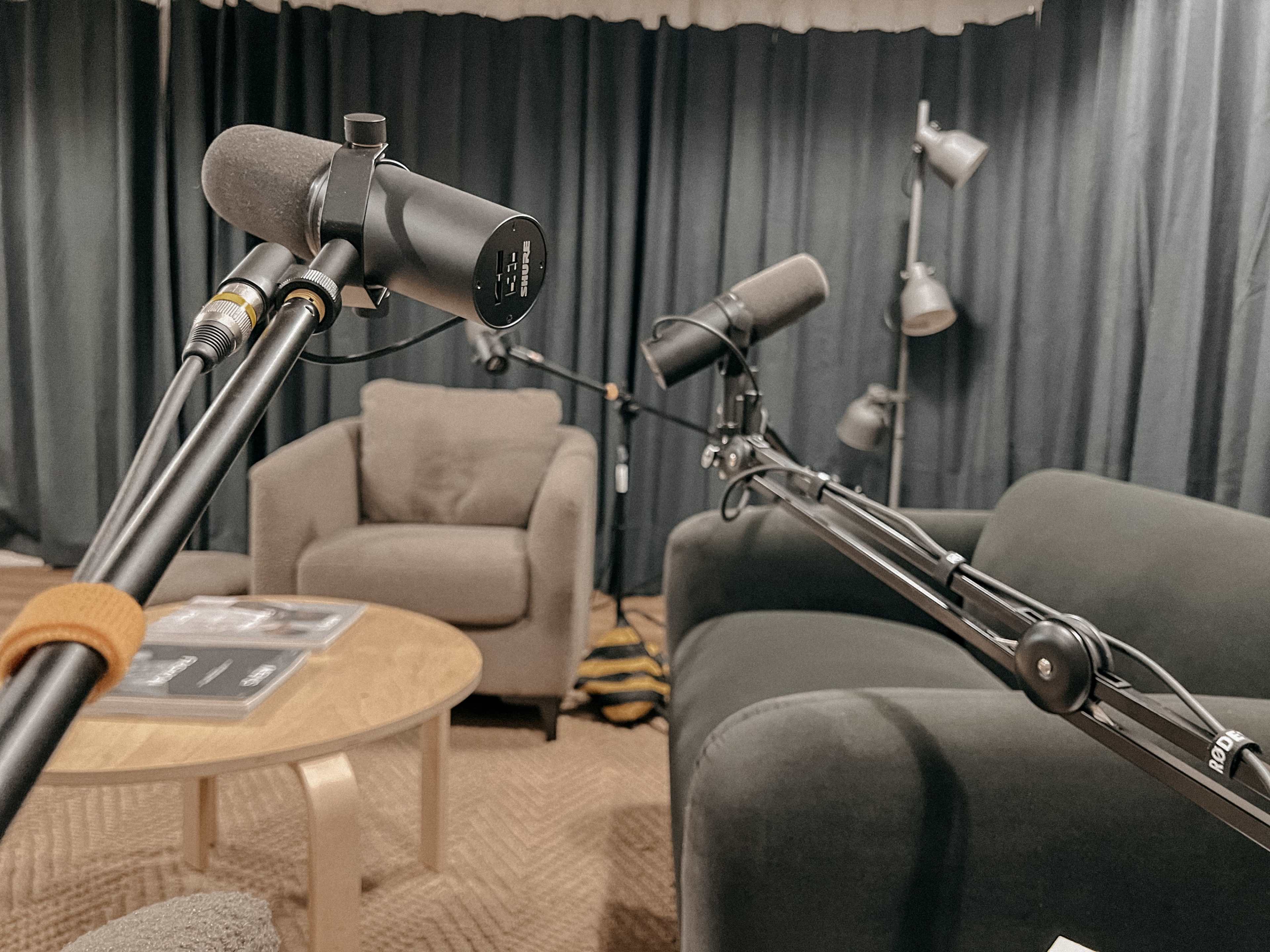 Two microphones on boom arms positioned near a coffee table and two chairs in a room with dark curtains.