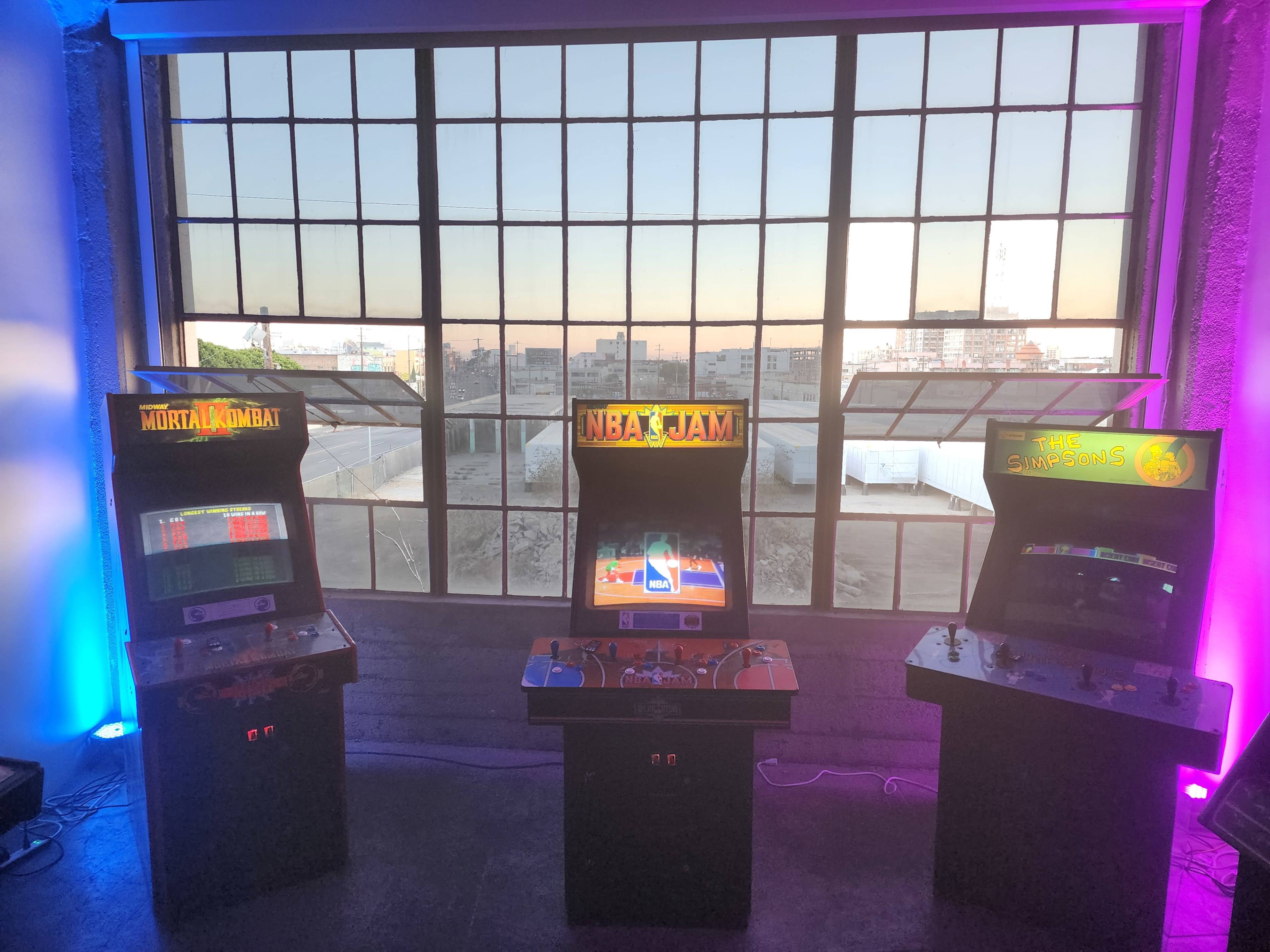 Three retro arcade machines, featuring "Mortal Kombat," "NBA Jam," and "Gauntlet," are positioned in front of a large industrial window with a city view.