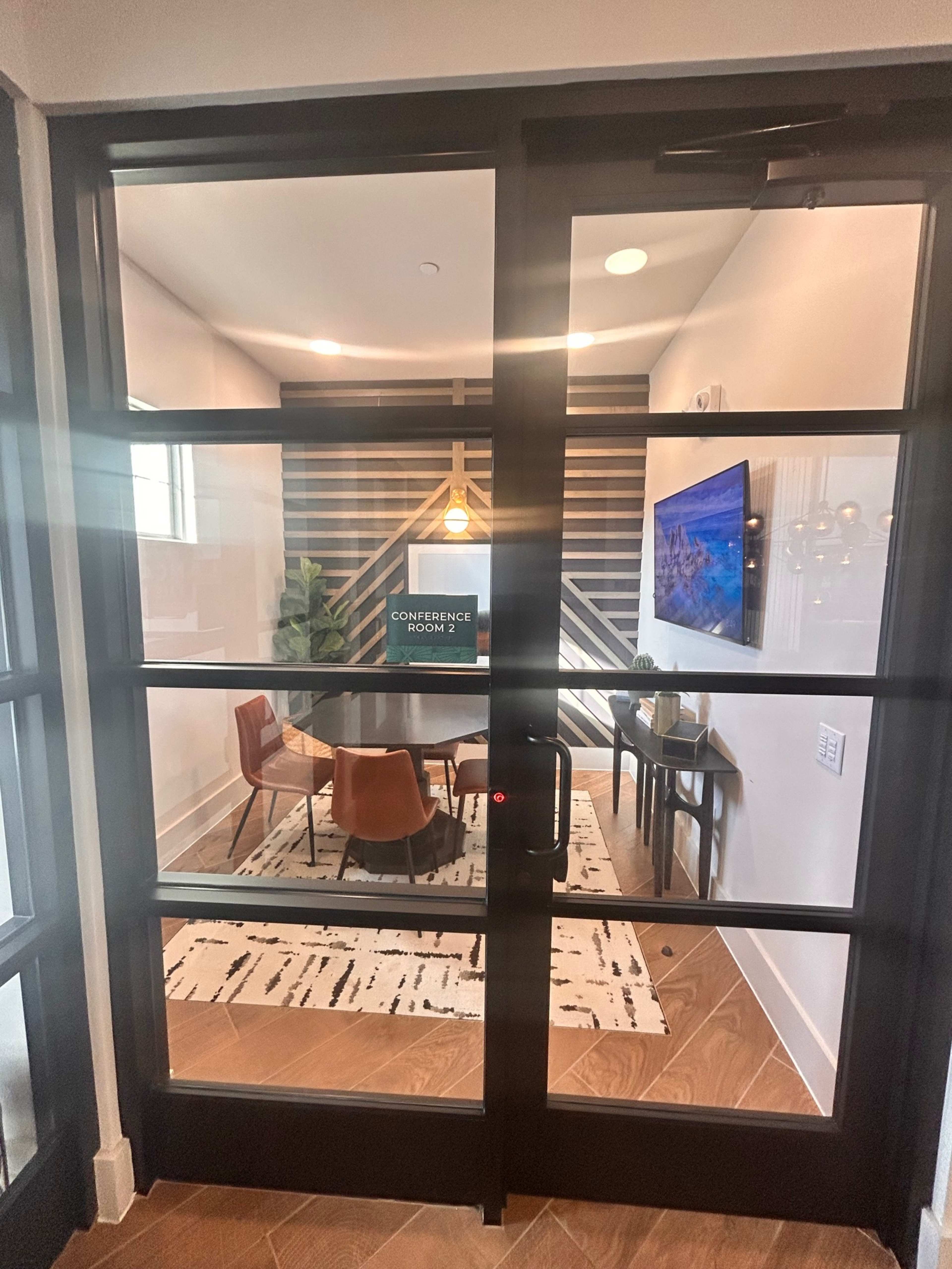 The image shows a glass door leading into a conference room featuring a round table with chairs, decorative plants, and a wall with a geometric design.