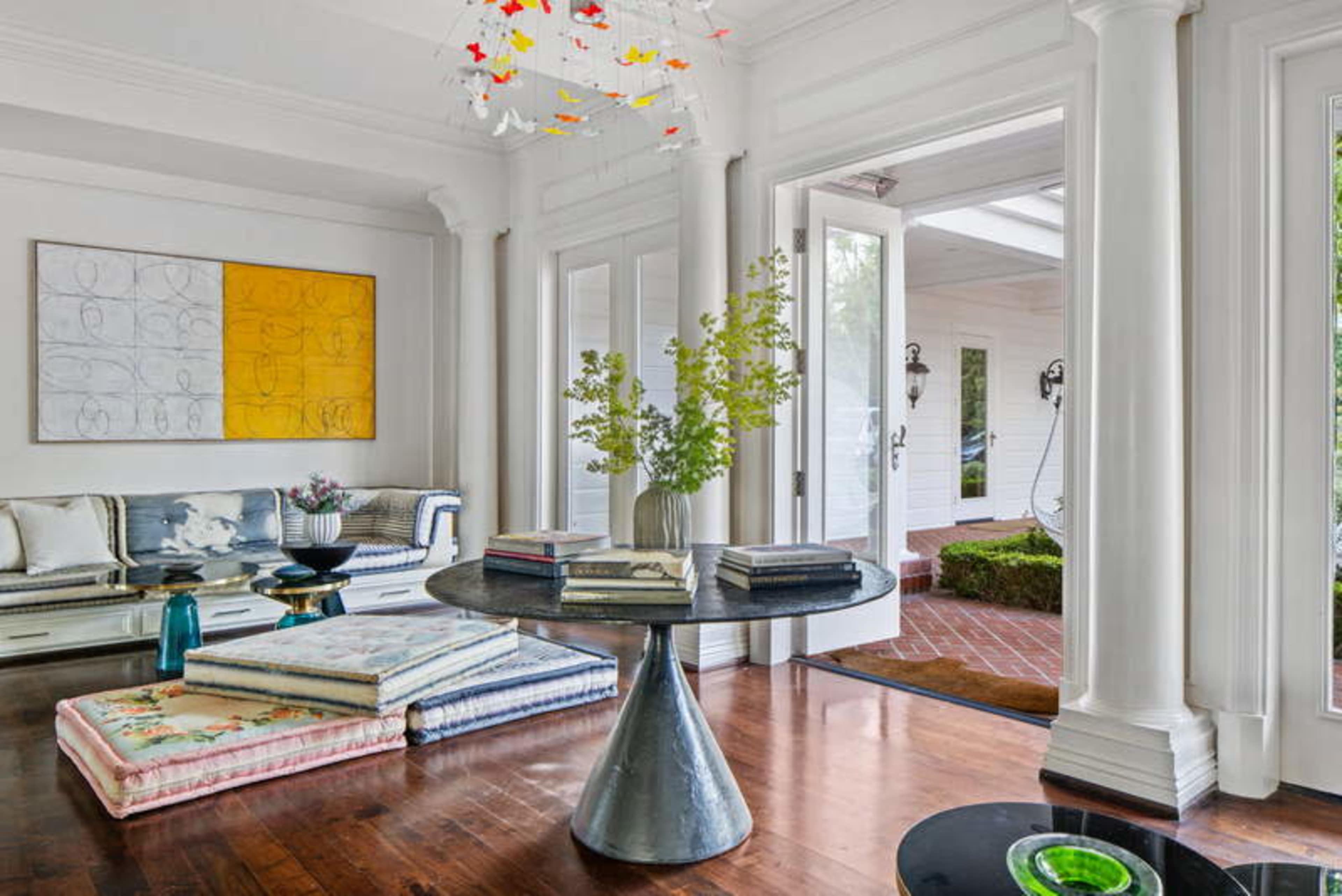 The image shows a bright room with a circular table at the center, surrounded by colorful seating and a door opening to an outdoor area.