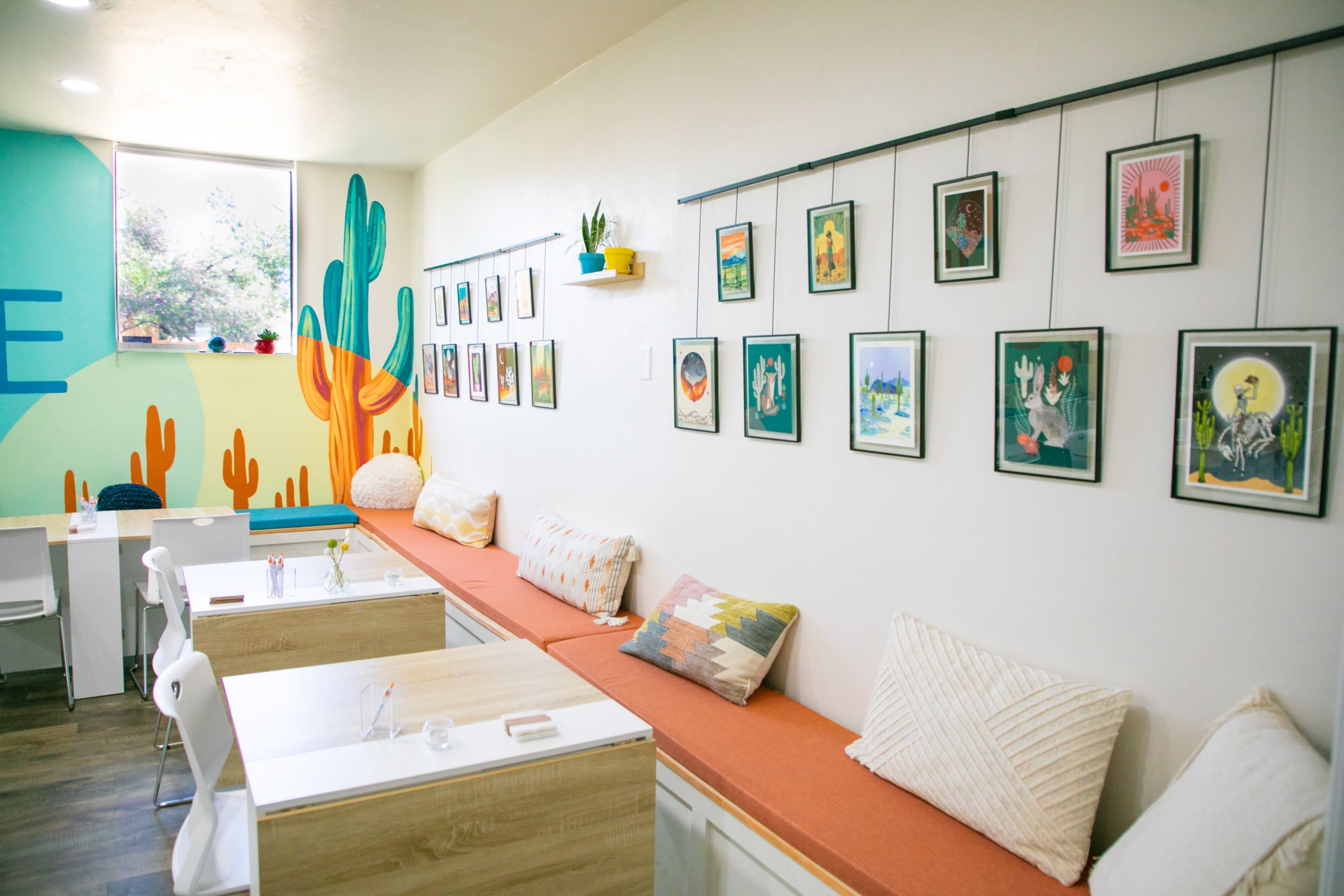 A bright café interior with a cozy seating area, decorated with framed artwork and plants against a colorful wall featuring cacti.
