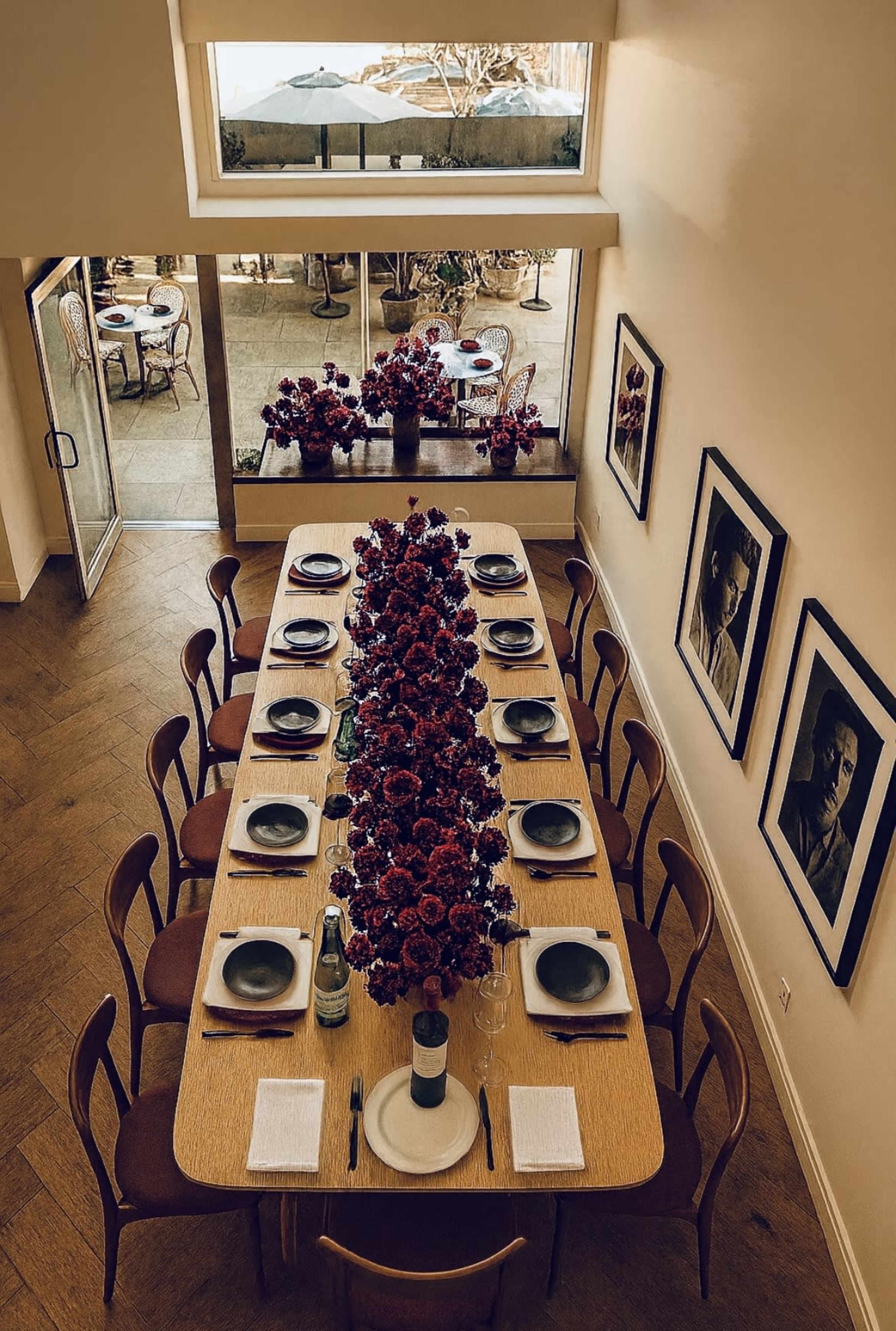 A long dining table is set with plates and glasses, flanked by floral arrangements, against a backdrop of framed photographs and large windows.