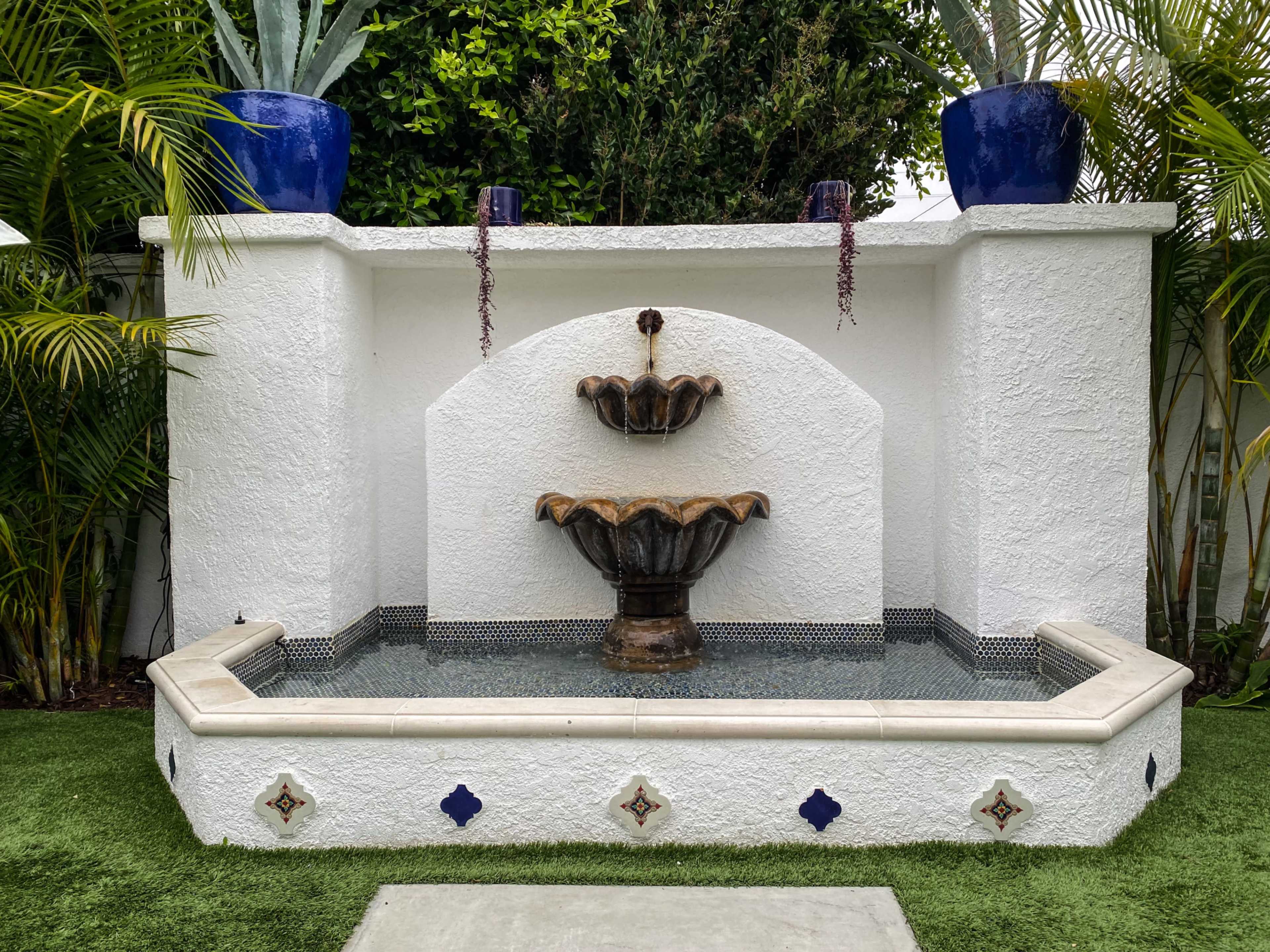 A two-tiered fountain set within a white stucco wall, surrounded by greenery and potted plants.